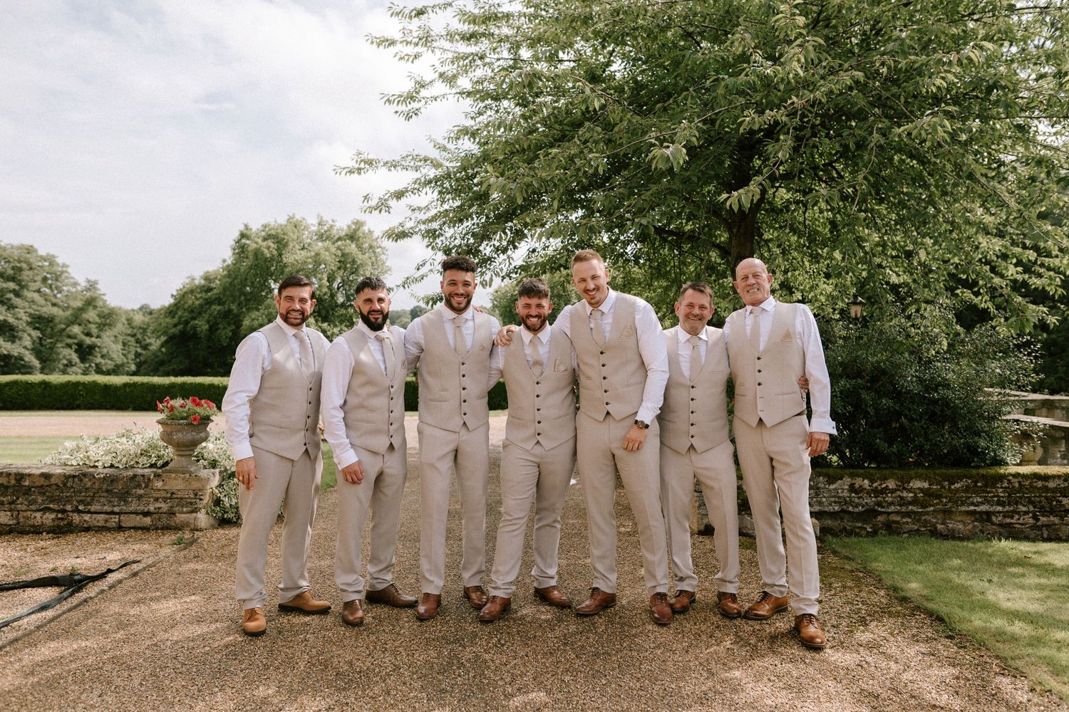 A group of groomsmen wearing matching beige suits and brown shoes pose for a photo in a garden setting.