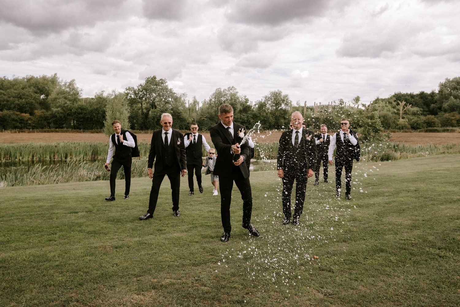 A group in formal black attire walking across a grass field under cloudy skies on a wedding day.