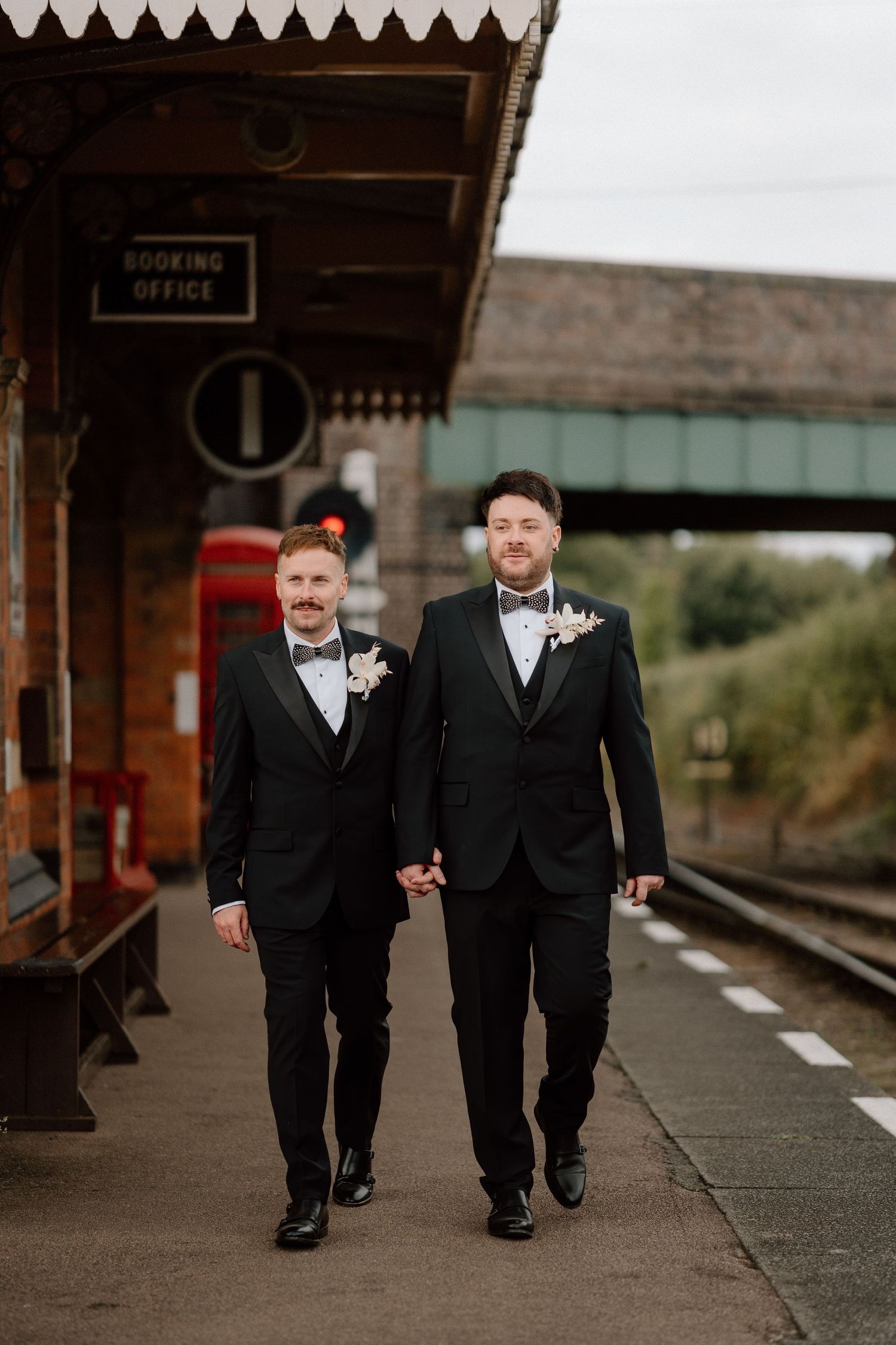 Two people in black tuxedos walking along a vintage train platform at The Manor House at Quorn.