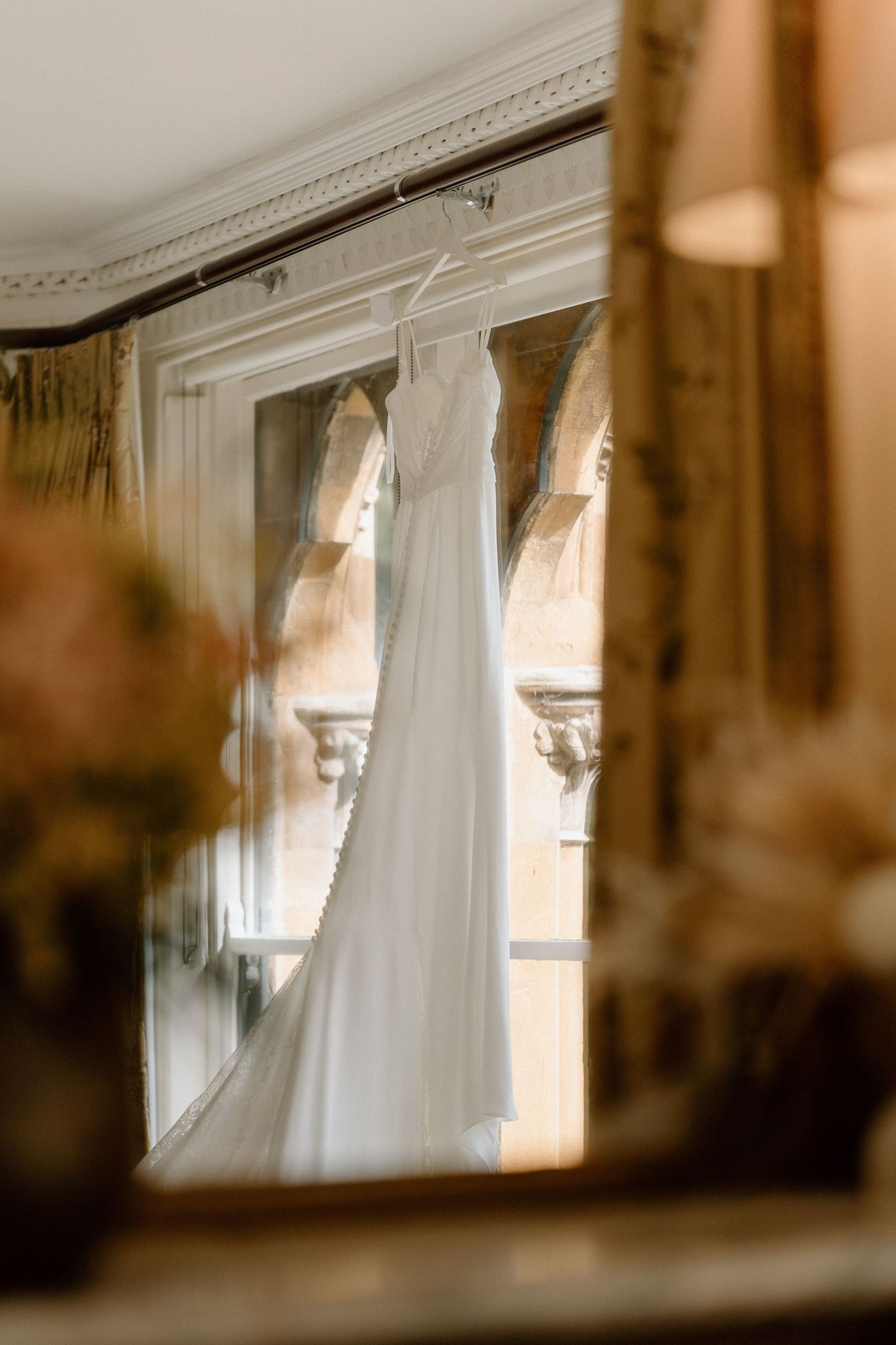 A wedding dress hanging in front of ornate windows, captured with soft natural lighting at Ettington Park Hotel.