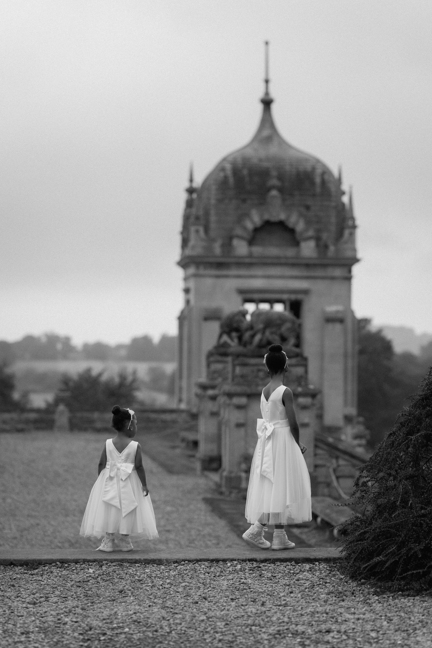 Two figures in white dresses stand before an ornate domed architectural structure in black and white at Harlaxton Manor.