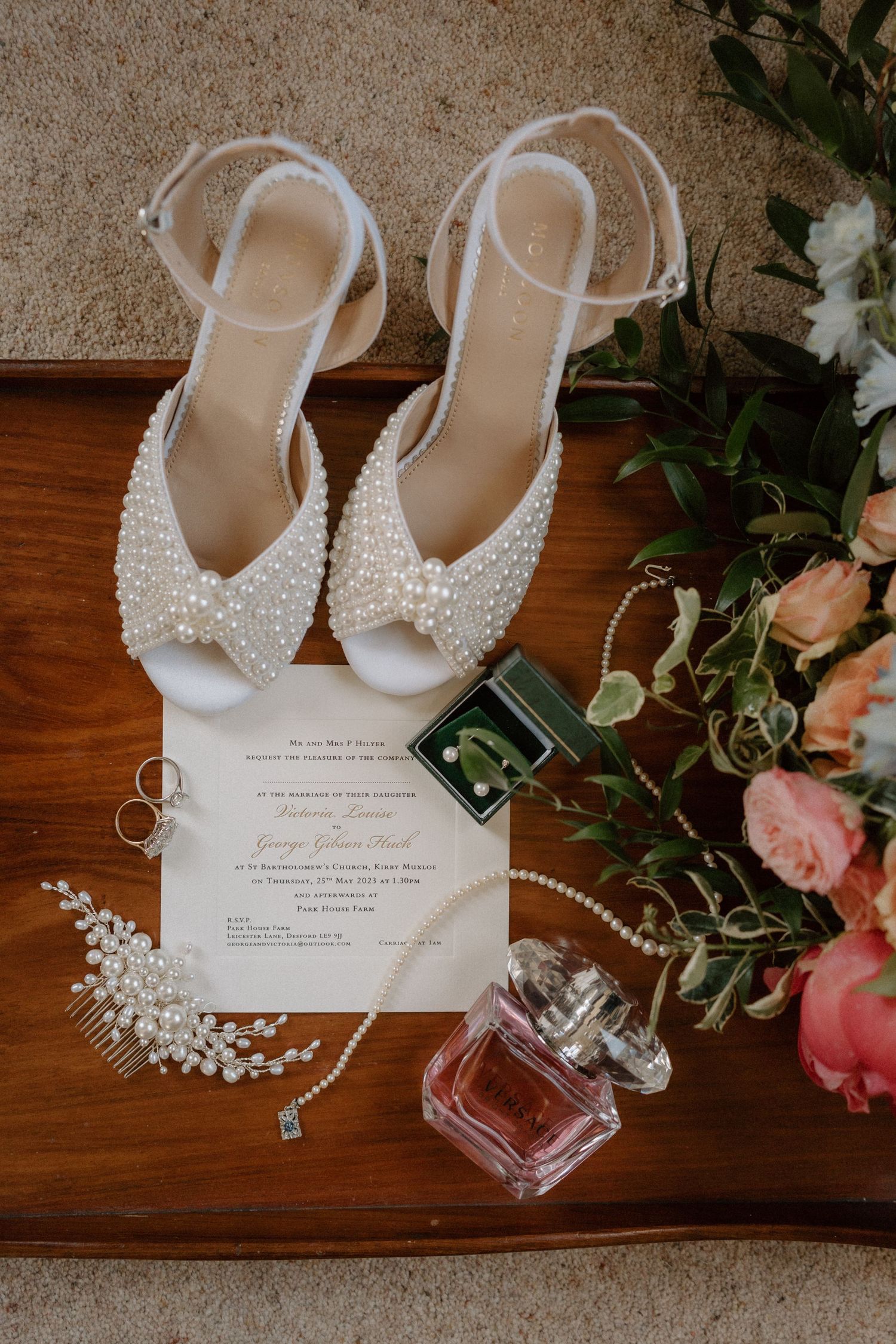 Wedding accessories including pearly shoes, invitation, jewelry and perfume arranged on wooden surface.