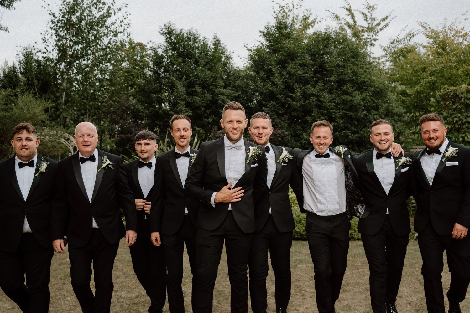 A group of groomsmen in black tuxedos and bowties stand together outdoors for a wedding photo at Warwick House.