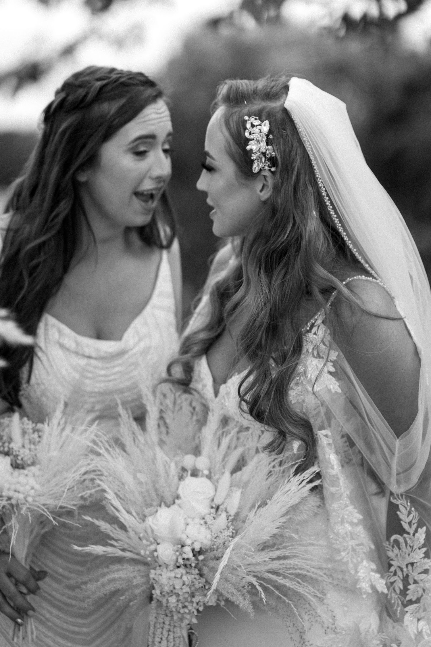 Two elegant figures share a candid moment while holding dried pampas grass bouquets at a wedding.