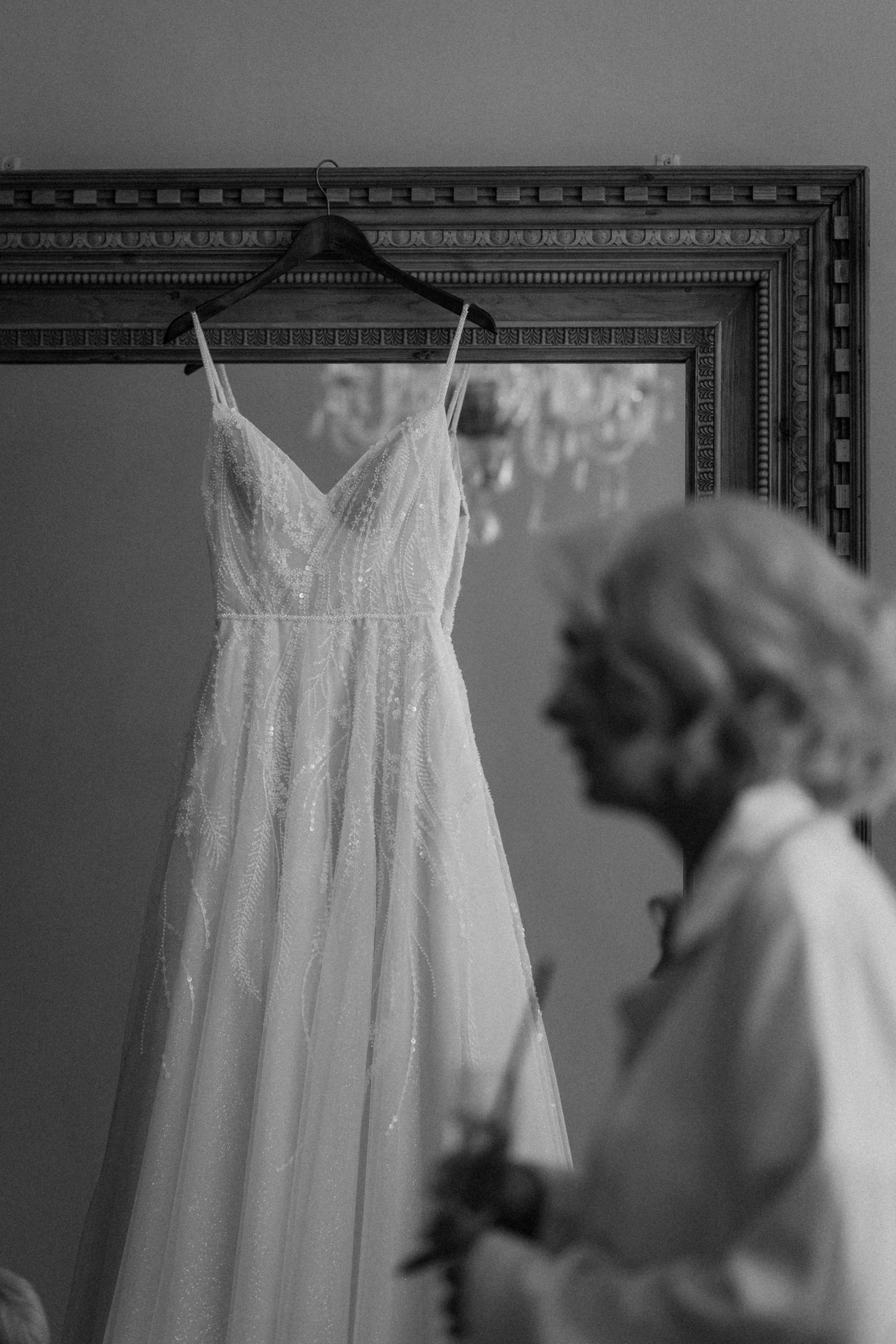 Black and white photo of an elegant A-line wedding dress hanging on a frame with a blurred woman in the background.