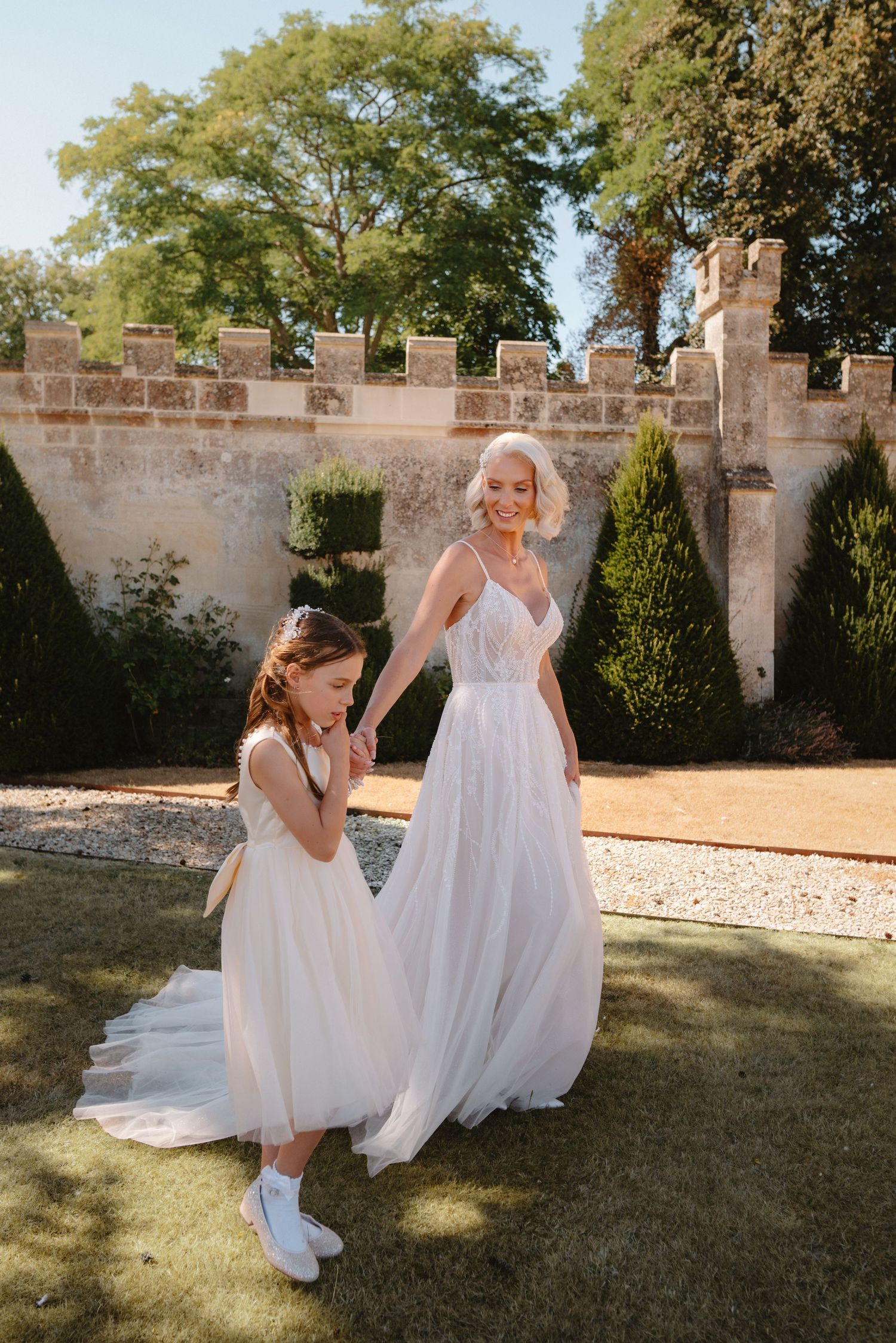 Bride in white tulle gown holds hands with flower girl in castle garden on sunny wedding day.
