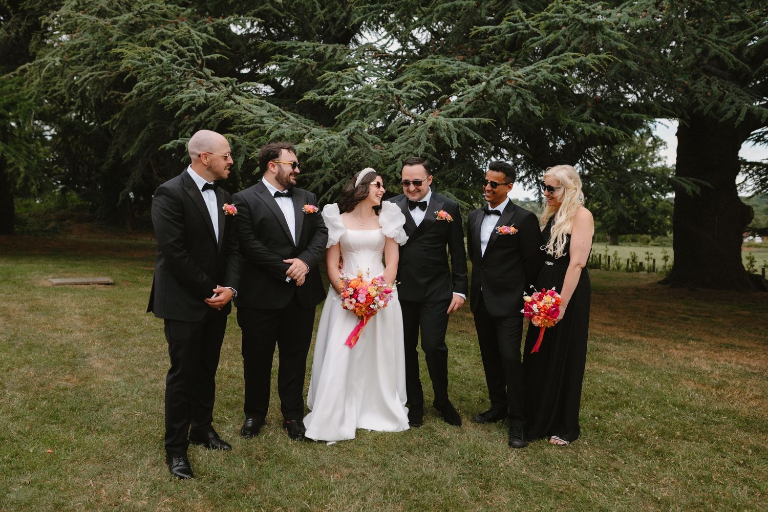 Wedding party posing outdoors, bride in white gown with groomsmen in black tuxedos and bridesmaids holding coral bouquets.