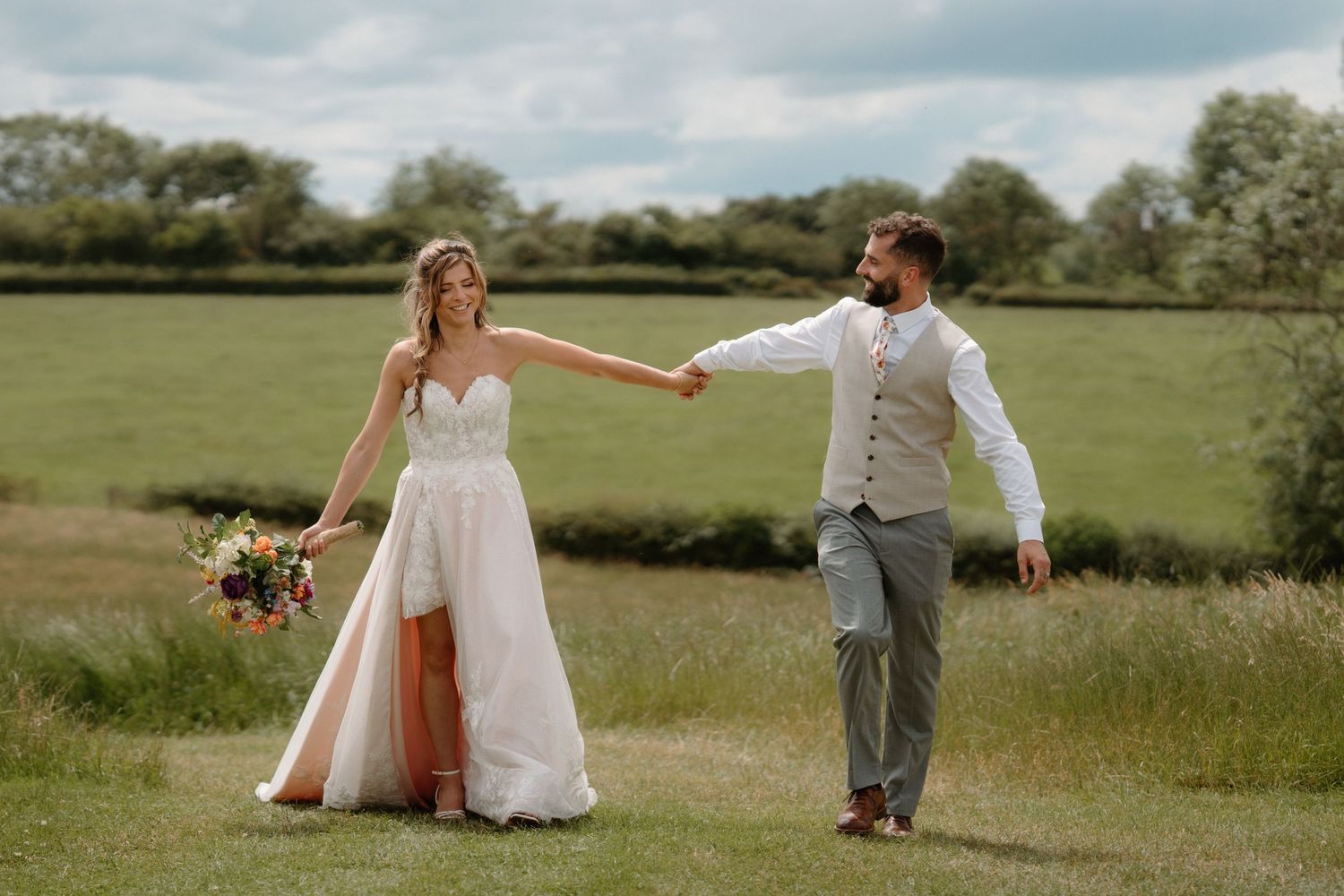 Bride and groom holding hands walking through green meadow, bride in white lace gown holding colorful bouquet.