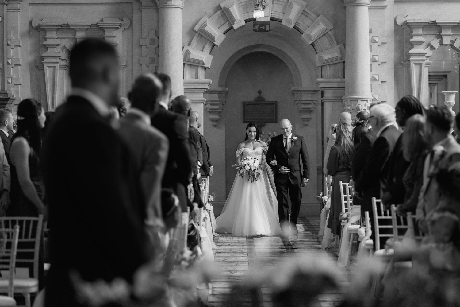A black and white ceremony photo showing guests seated in rows with stone archways in the background at Harlaxton Manor.
