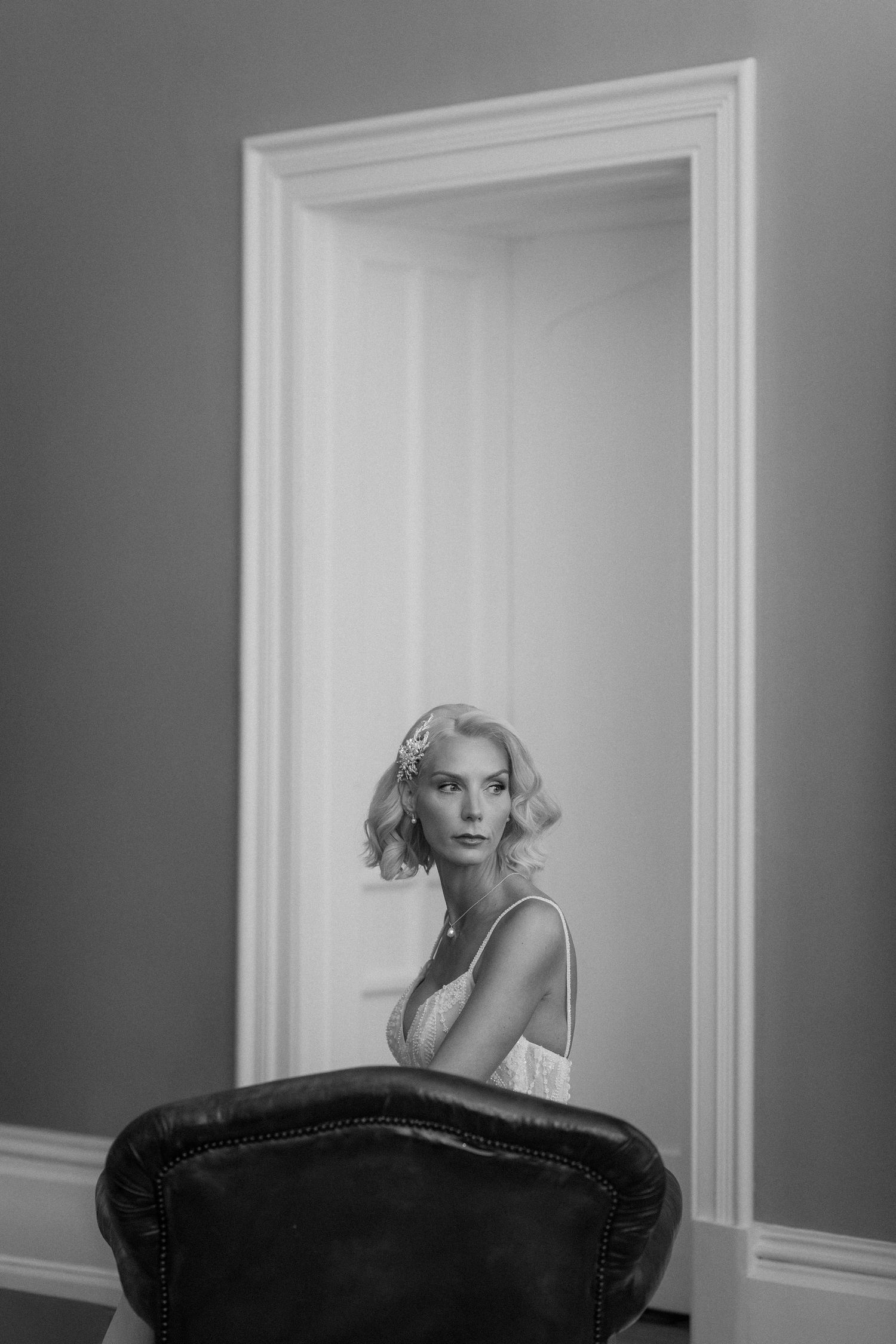 Black and white portrait of an elegant blonde woman in a doorway, wearing a white dress with floral hair accessory.