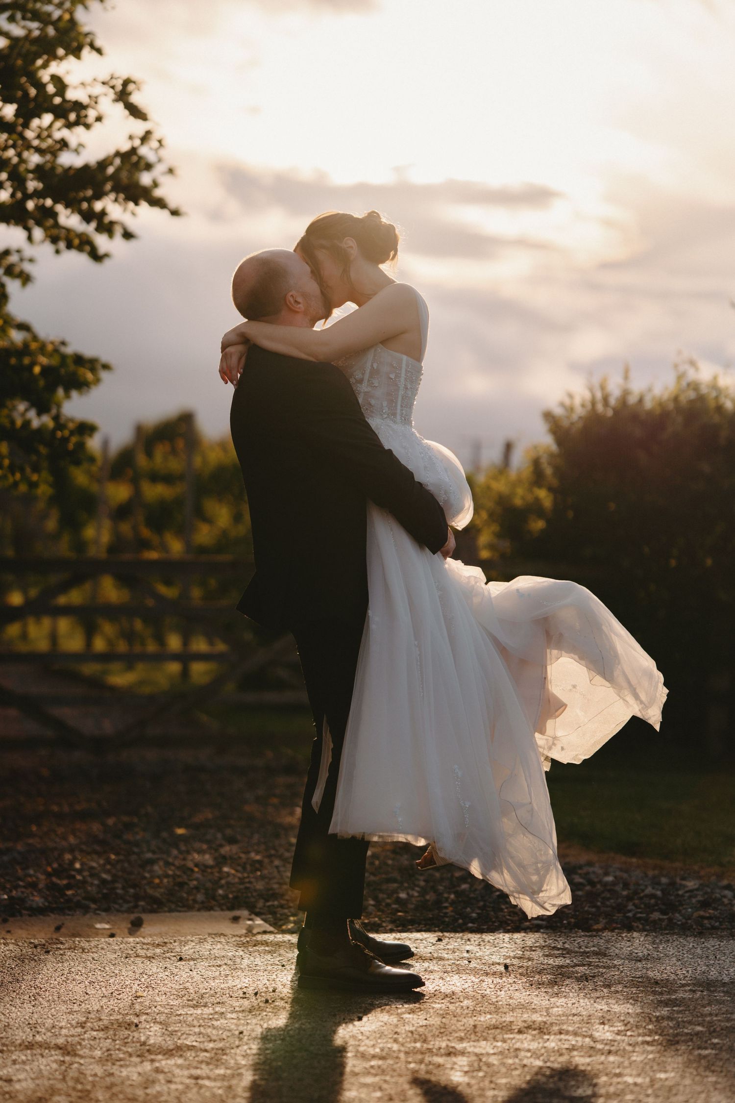 Groom lifting bride in white gown at golden hour, sharing a romantic kiss outdoors with soft sunlight behind them.