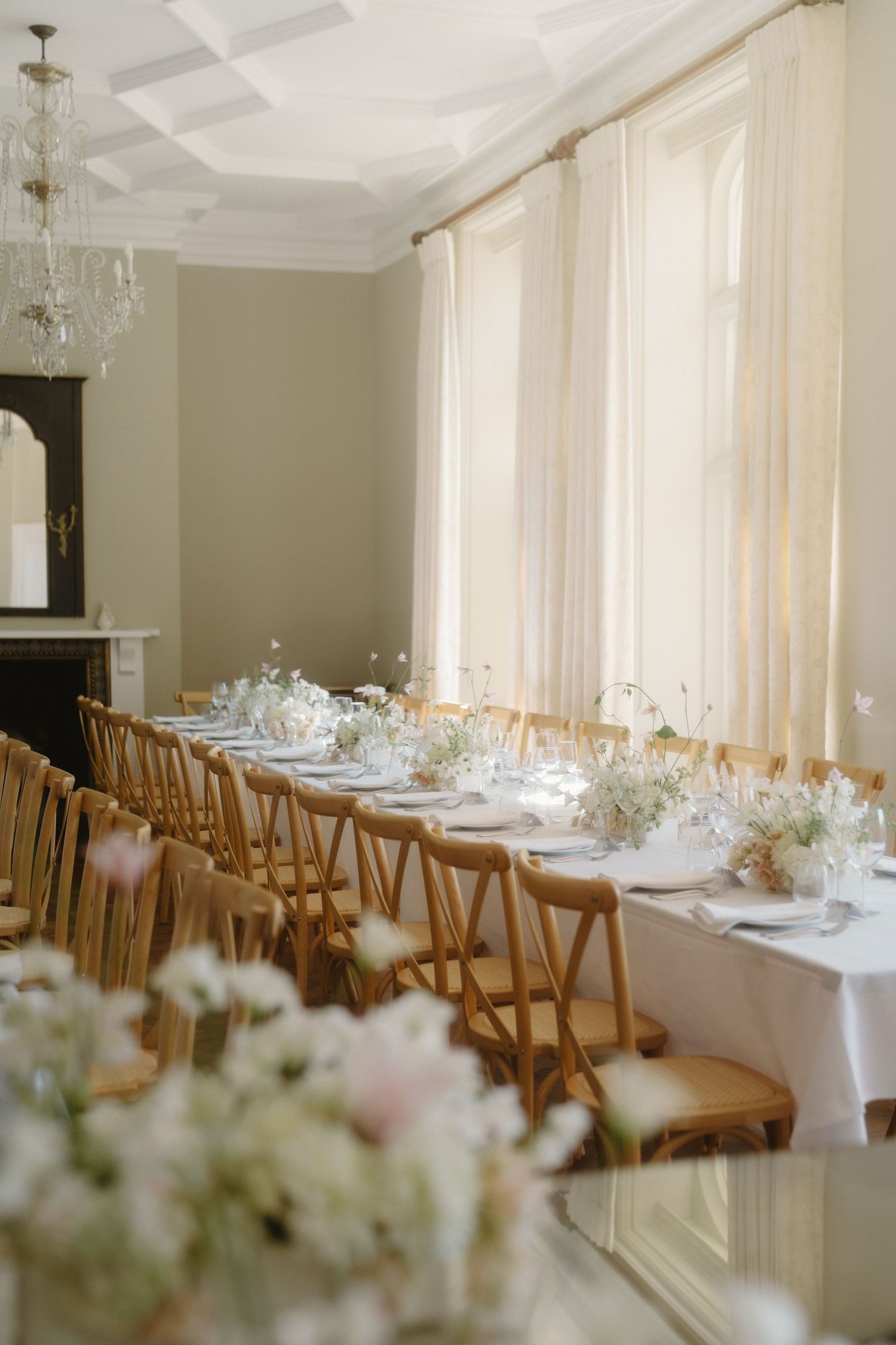 Elegant wedding reception with long table, wooden cross-back chairs, white floral centerpieces, and sheer curtains in a classic room.