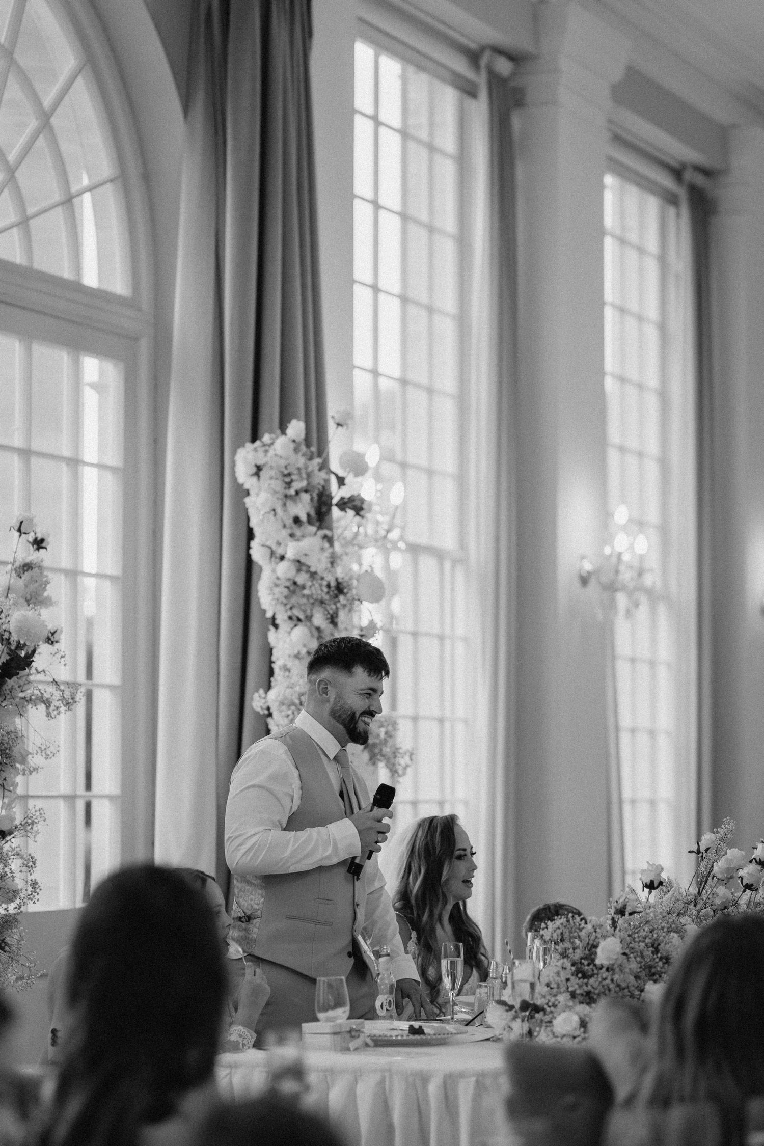 Black and white photo of a wedding reception speech being delivered in a bright room with tall windows.