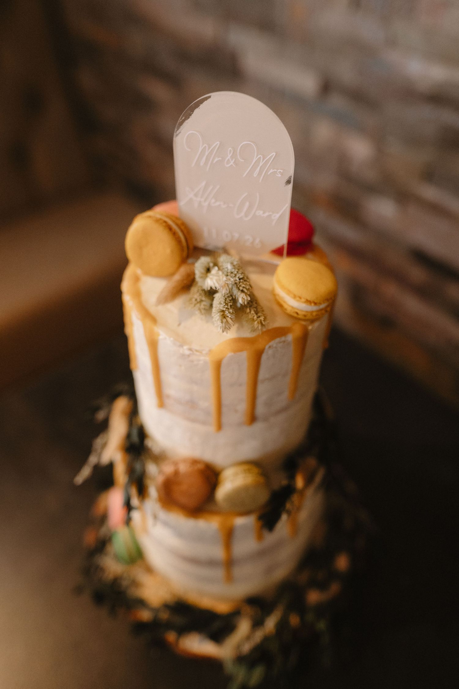 Two-tier naked wedding cake with caramel drip, macarons, and a personalized Mr. & Mrs. topper on a rustic wooden base.