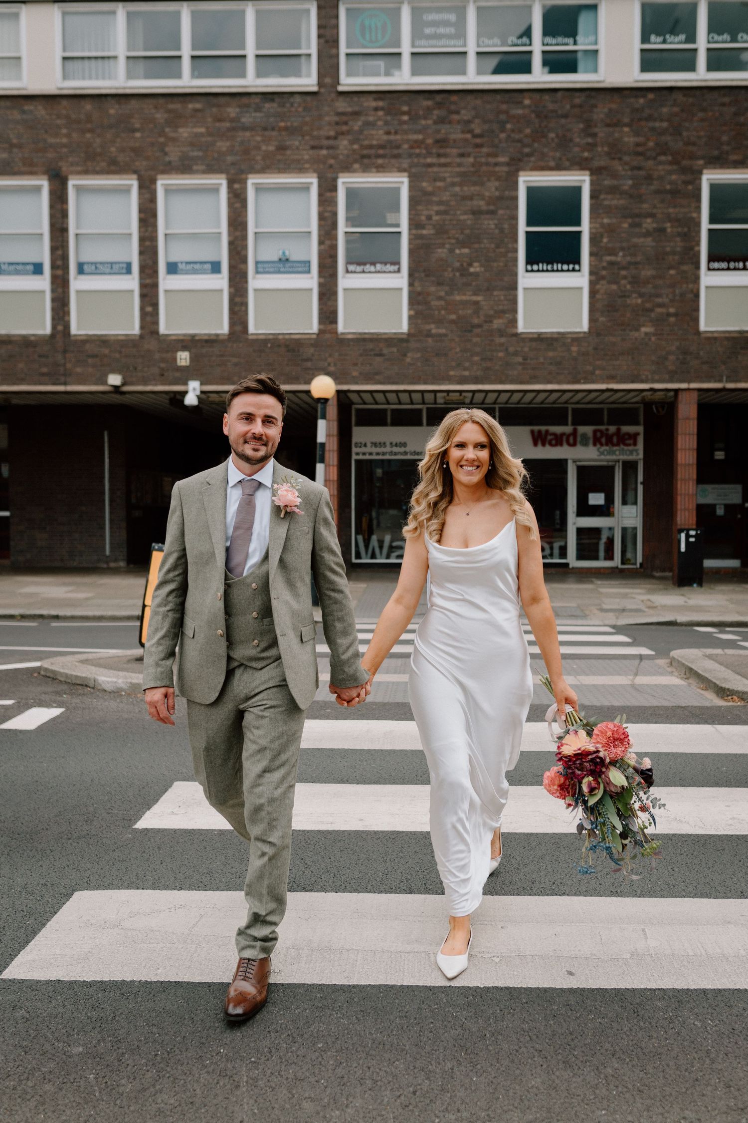 Couple in wedding attire walks hand in hand across a crosswalk in an urban setting in Coventry.