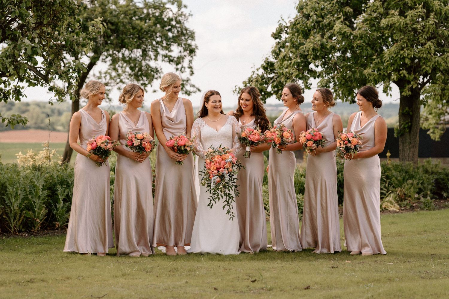 Wedding party in champagne coloured dresses posing together outdoors on a sunny day.