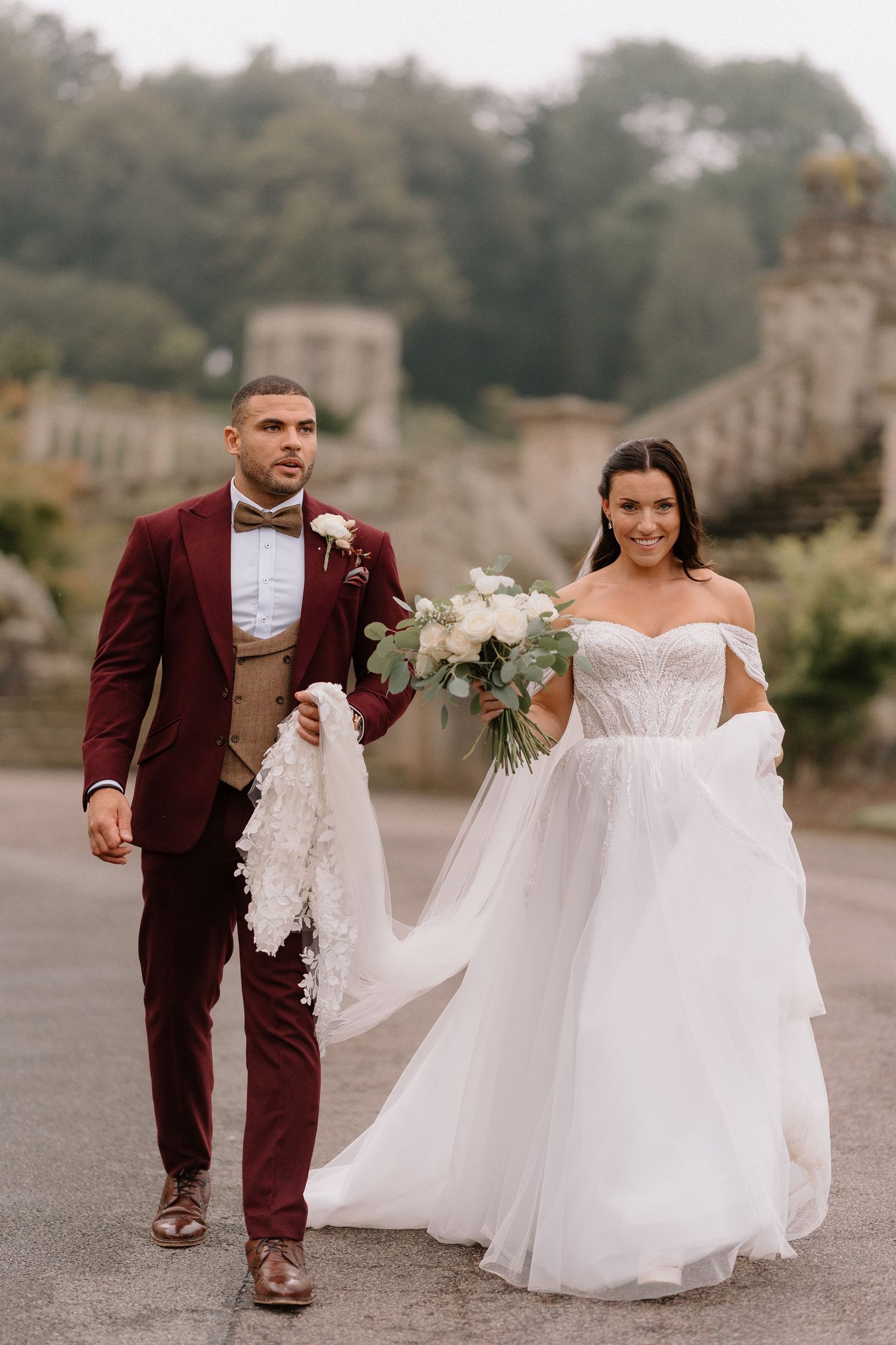 A wedding couple walks together on castle grounds, with the groom in burgundy and bride in flowing white gown at Harlaxton Manor.