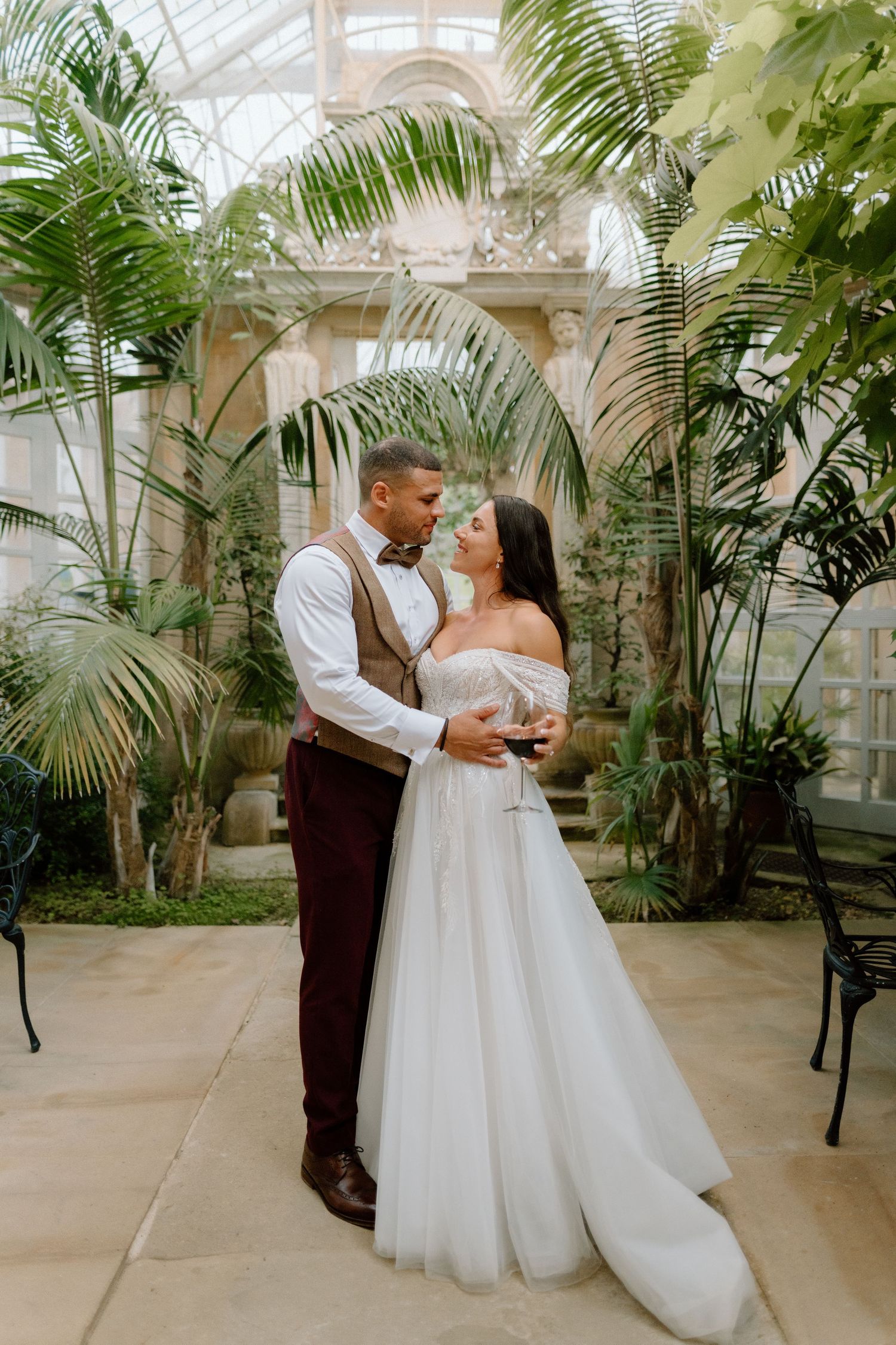 A romantic wedding photo in a greenhouse conservatory with palm trees and stone archways at Harlaxton Manor.