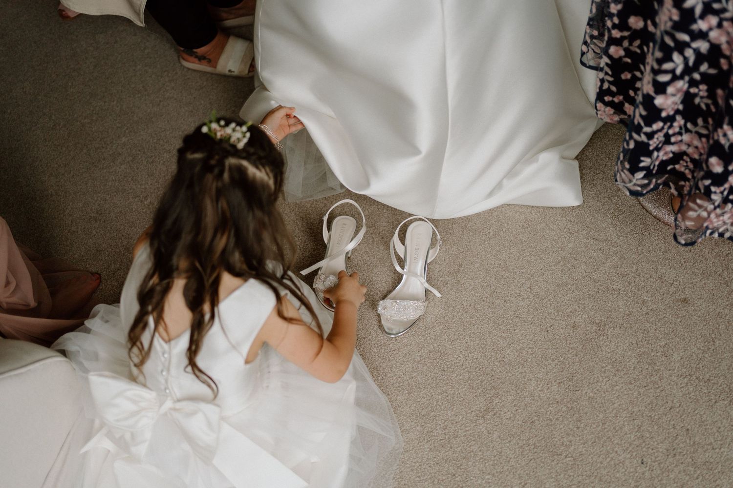 A little girl in a white dress adjusts the hem of a wedding dress while sitting on beige carpet at Dodford Manor.