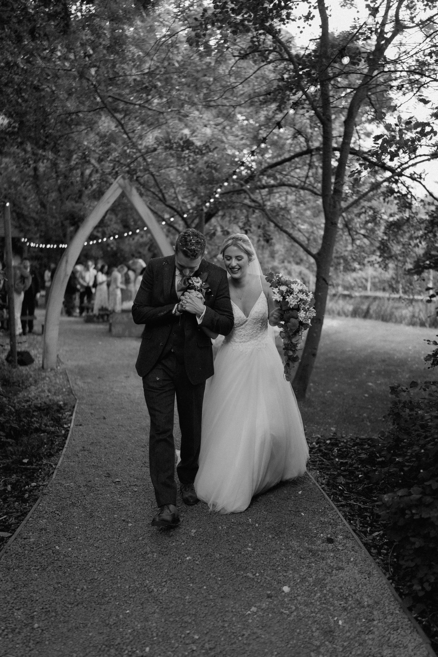 Black and white photo of couple walking under string lights on tree-lined path at Hothorpe Hall.