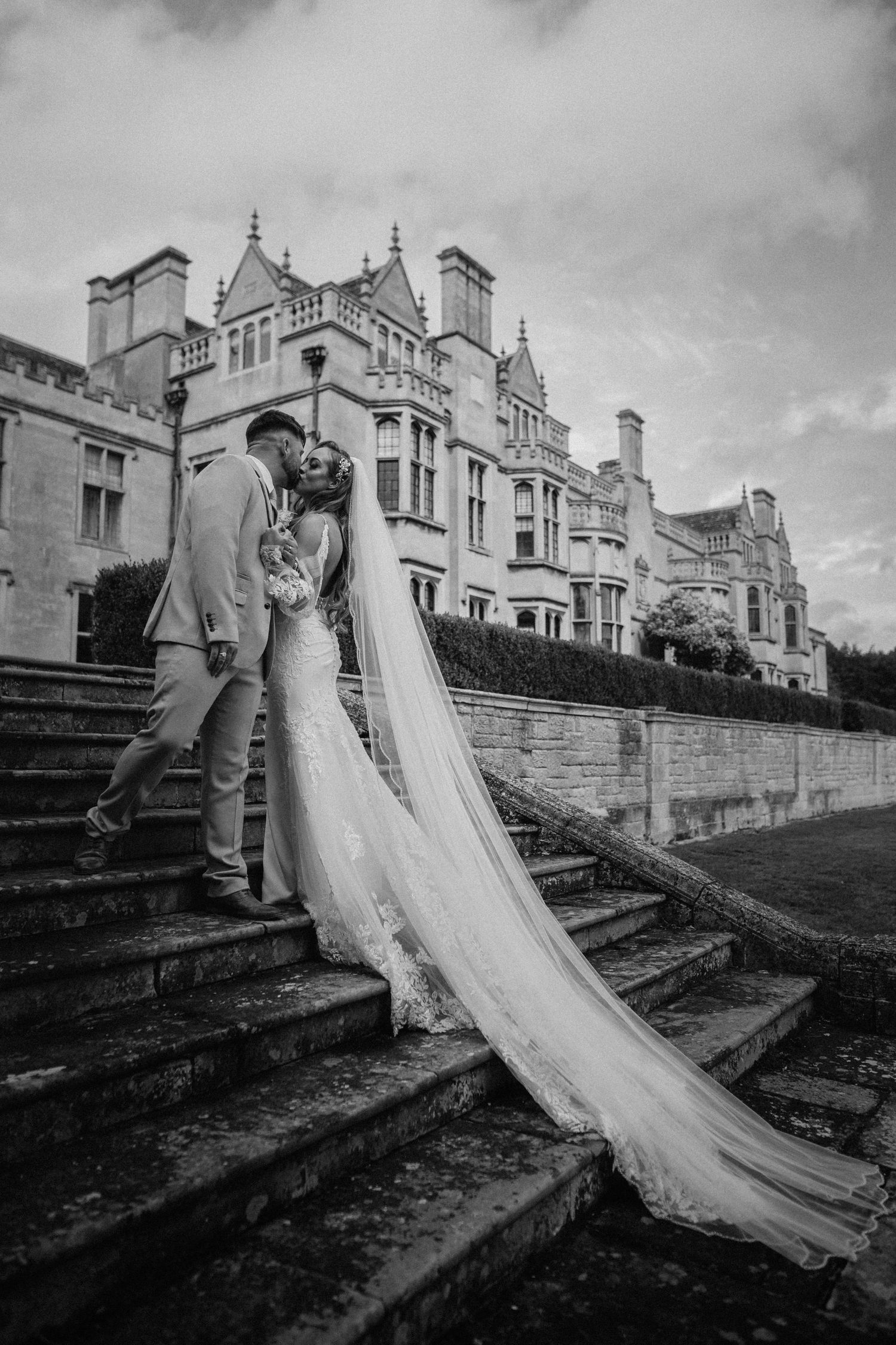 Black and white series of wedding couple on grand stone steps with Gothic mansion backdrop. Wedding photography at Rushton Hall. Northampton