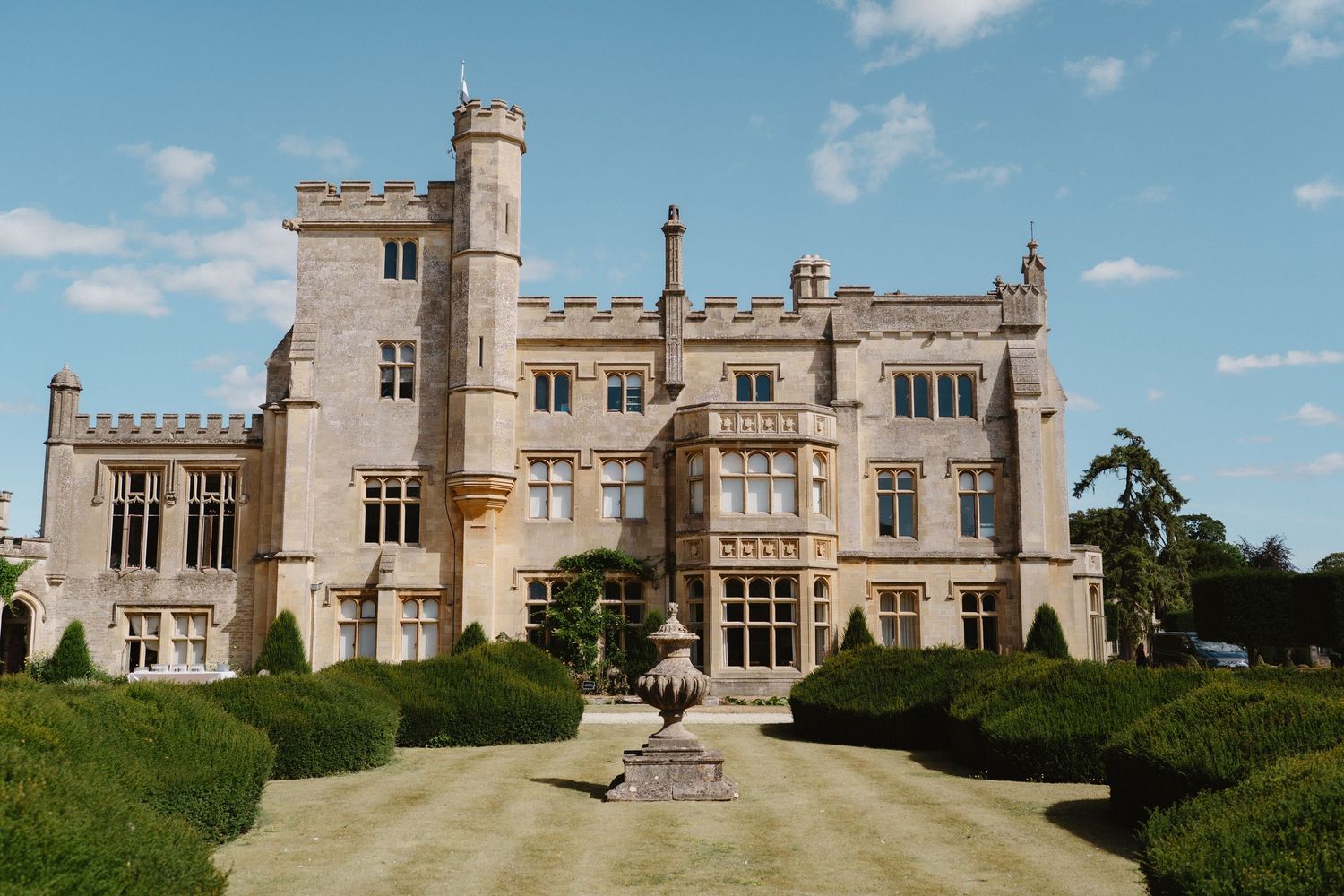 Grand English manor house with castellated tower, ornate stone facade, and manicured topiary gardens under a blue sky.