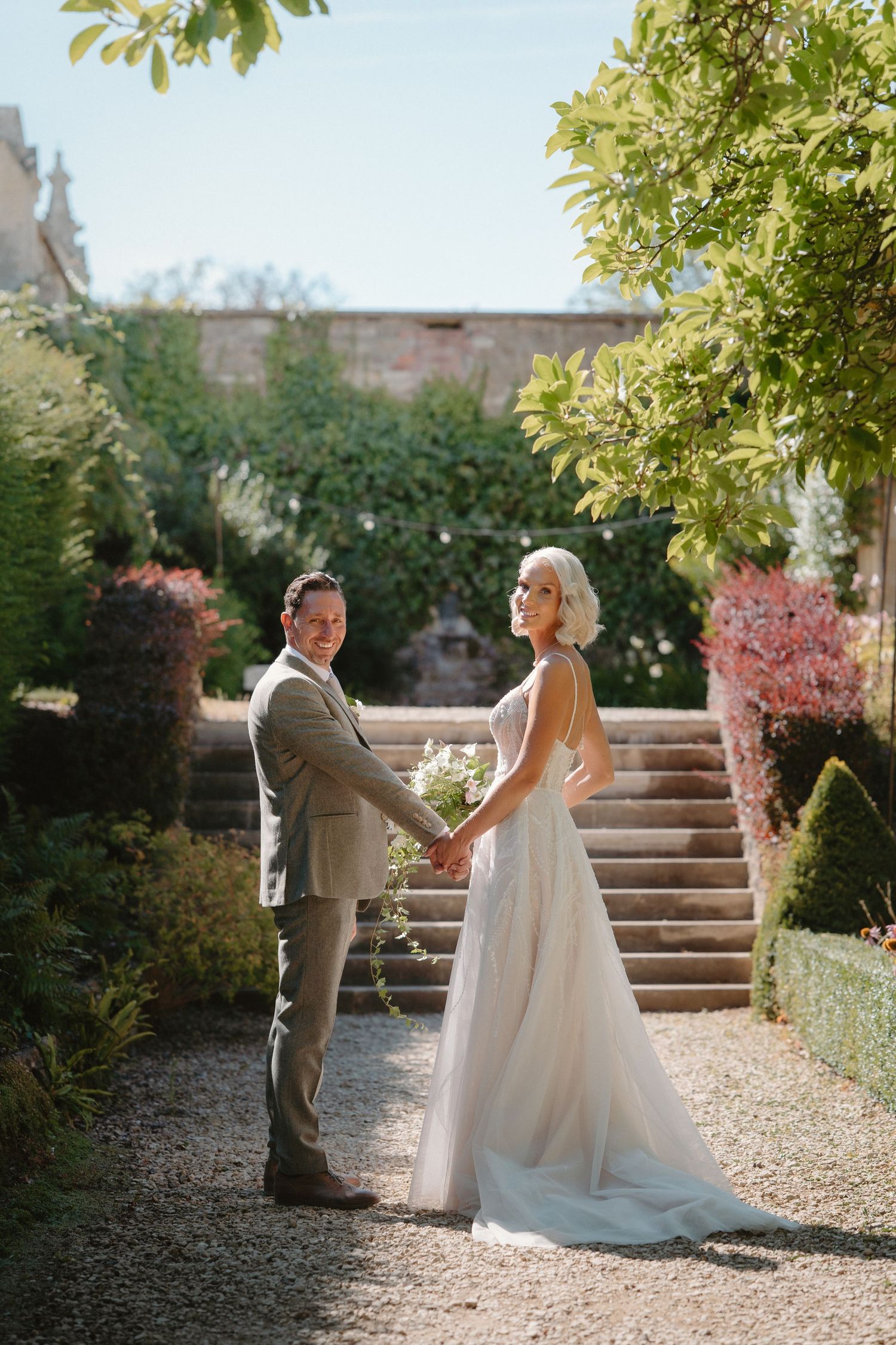 Bride and groom holding hands in a sunlit garden with stone steps and lush greenery on their wedding day.