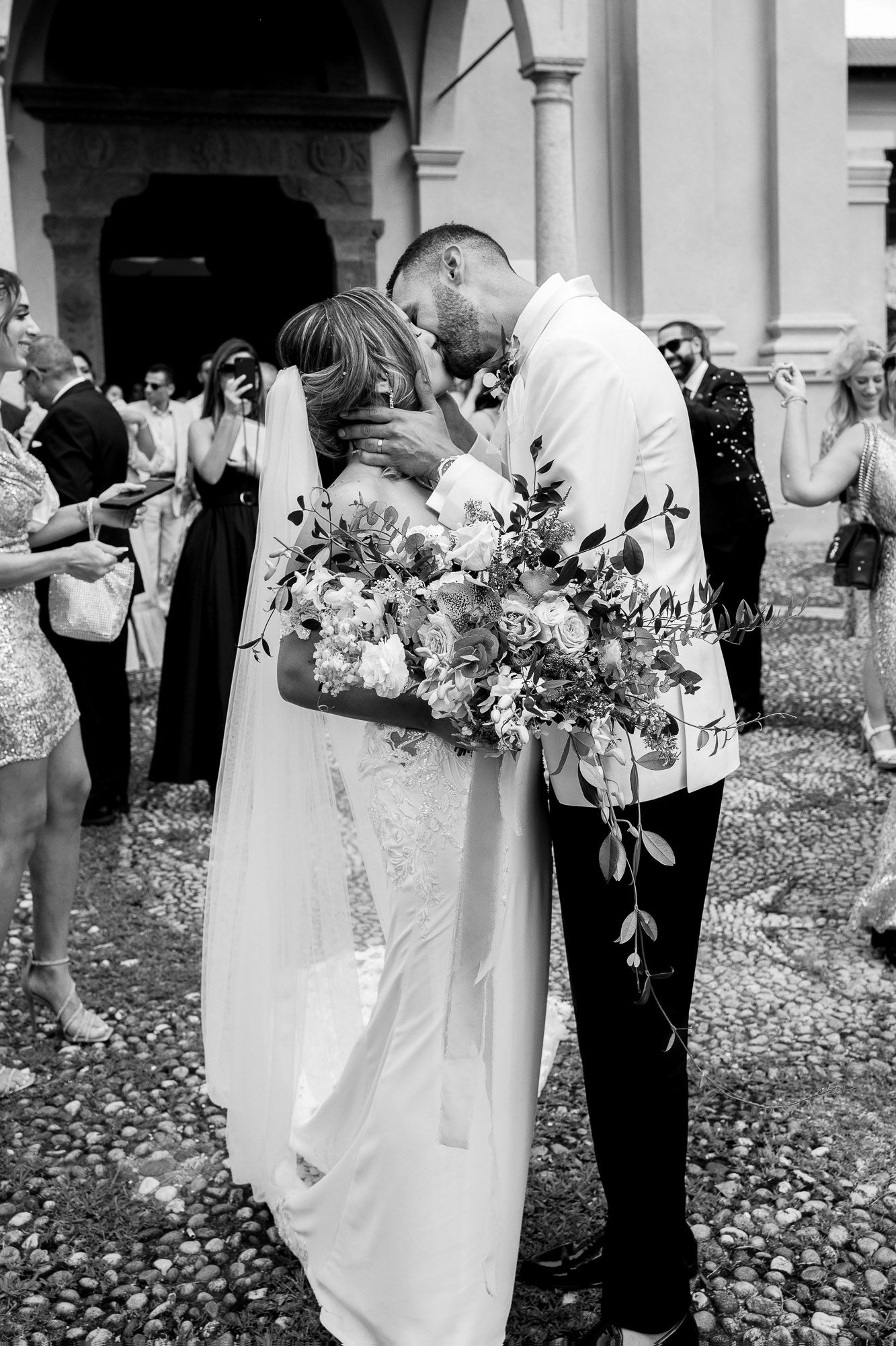 A romantic black and white wedding moment with the couple embracing while holding a beautiful floral bouquet outside a church.