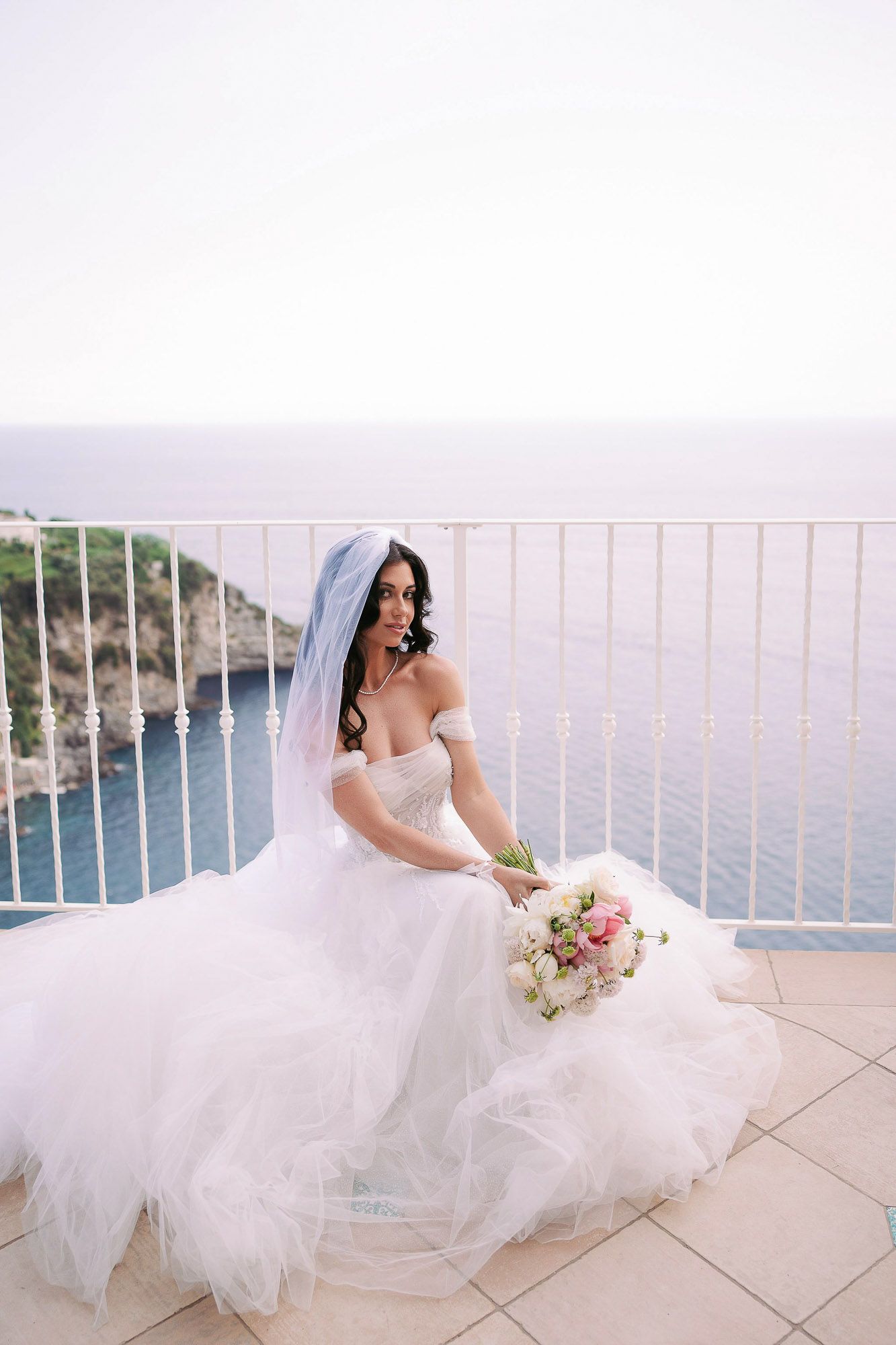 Bride in flowing white gown poses on seaside terrace overlooking coastal cliffs and blue waters.