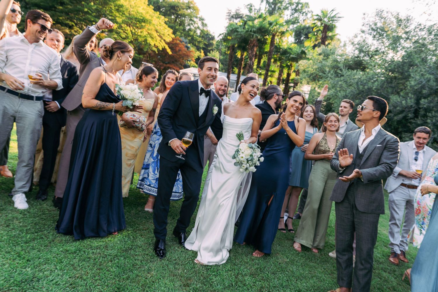 Wedding party gathered on a lush green lawn reacting with joy and excitement during an outdoor celebration.