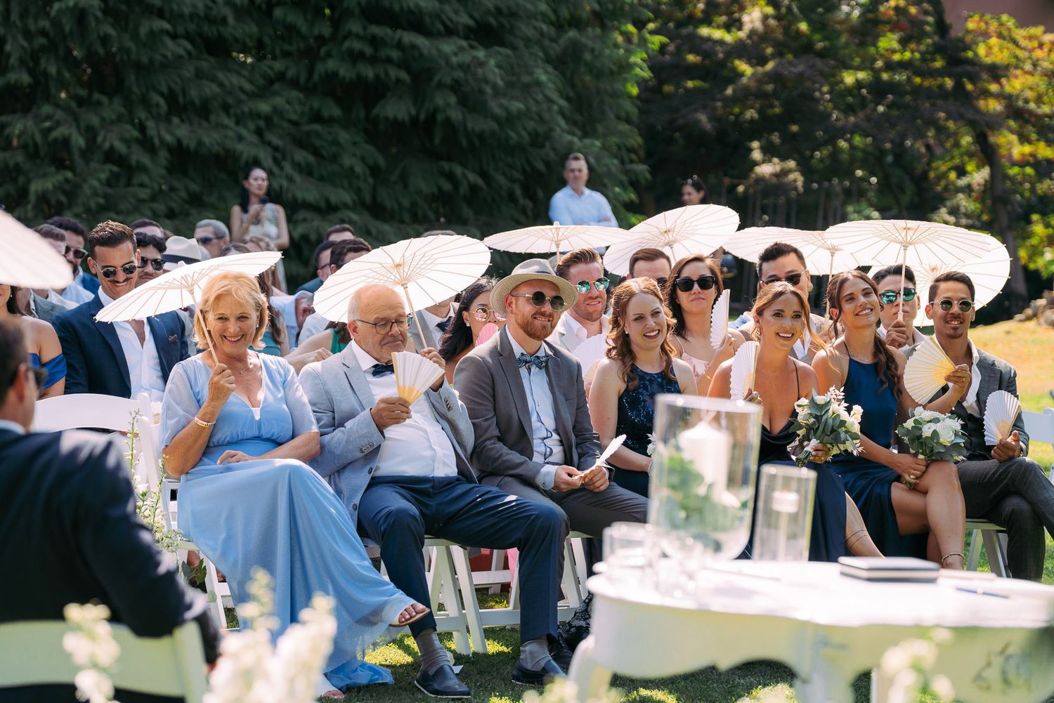 Wedding guests laugh and celebrate under white umbrellas during an outdoor garden reception on a sunny day.