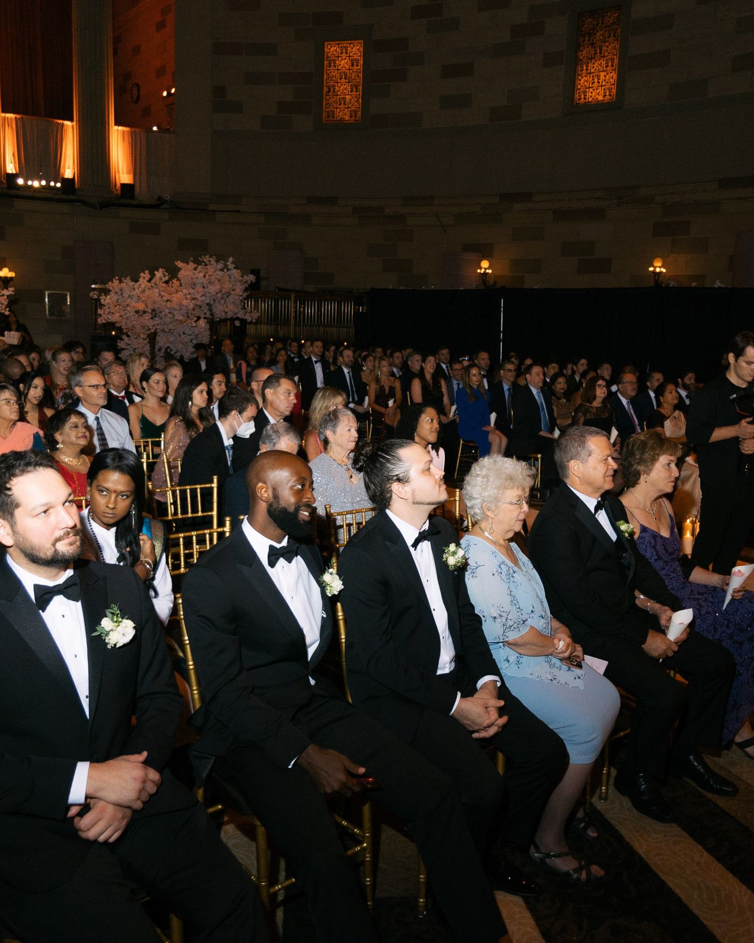 A formal evening event shows a crowded audience in black tie attire seated in rows at an elegant venue.