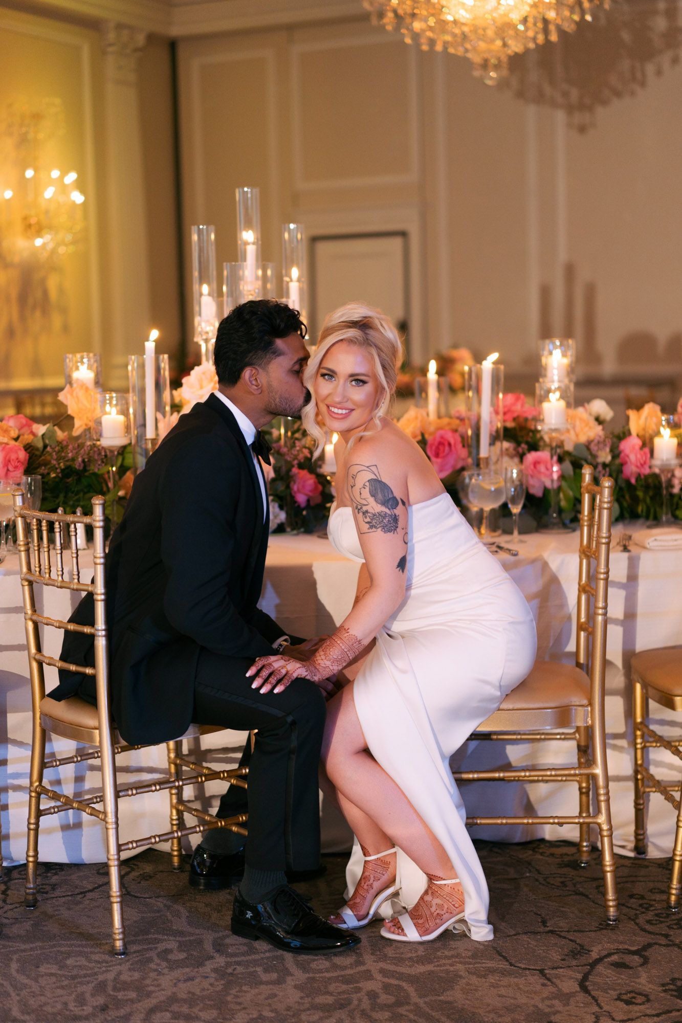 Wedding couple shares playful moment as groom removes bride's garter in elegant ballroom with crystal chandeliers.