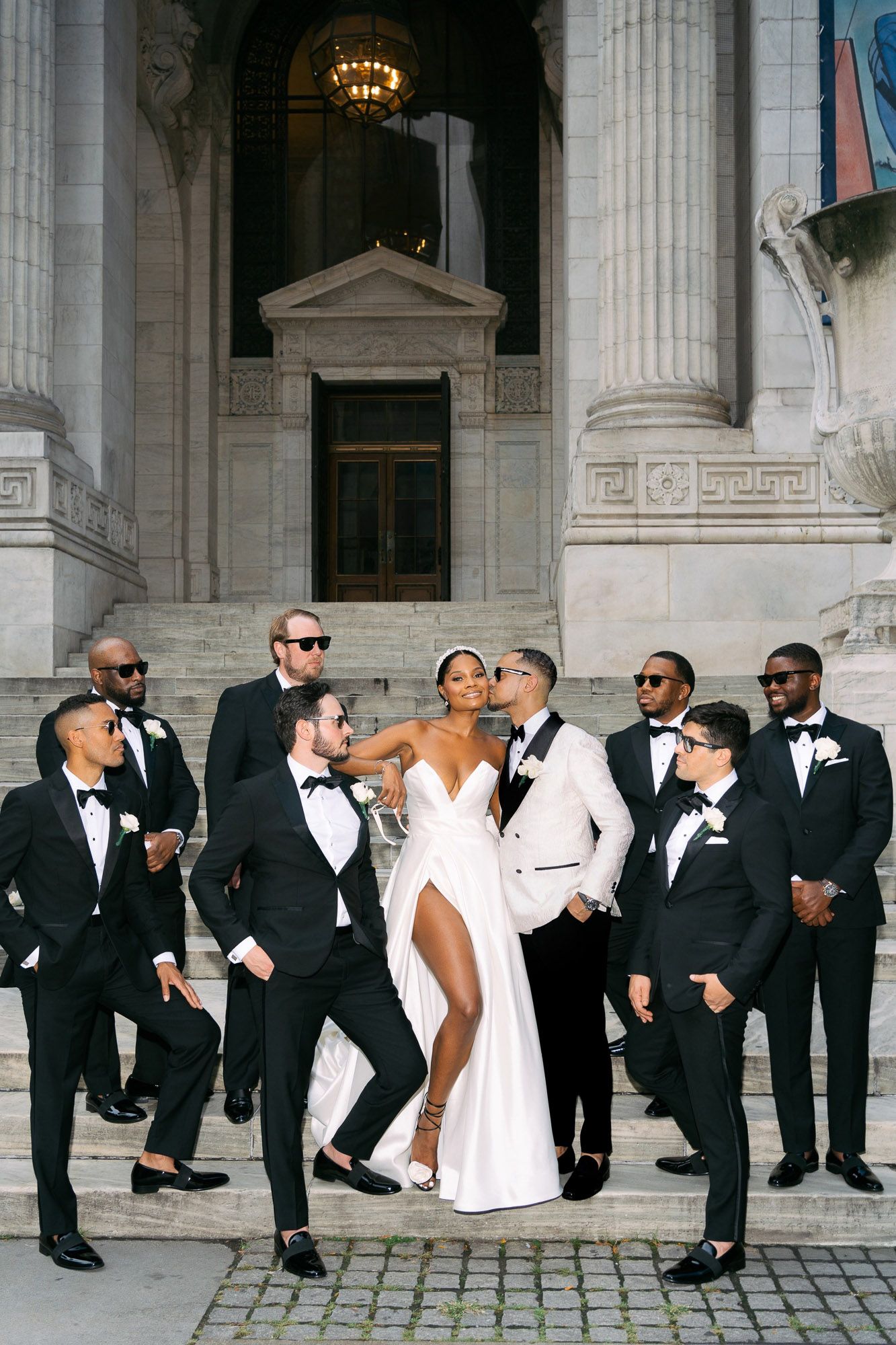 Wedding party in black and white formal attire poses creatively along columned building entrance.