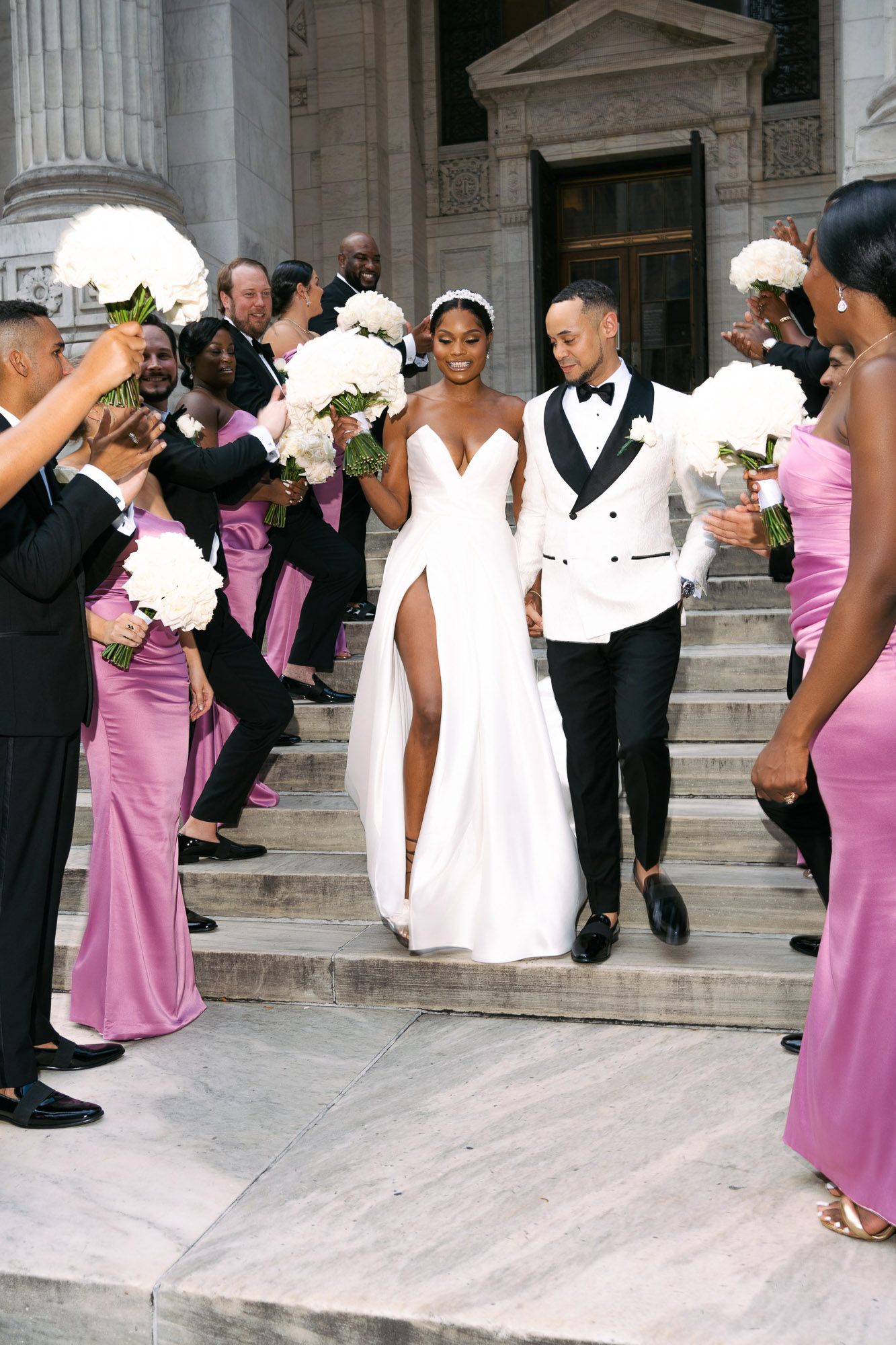 Newlyweds descend grand marble steps while wedding party in pink dresses and dark suits celebrate with white bouquets.