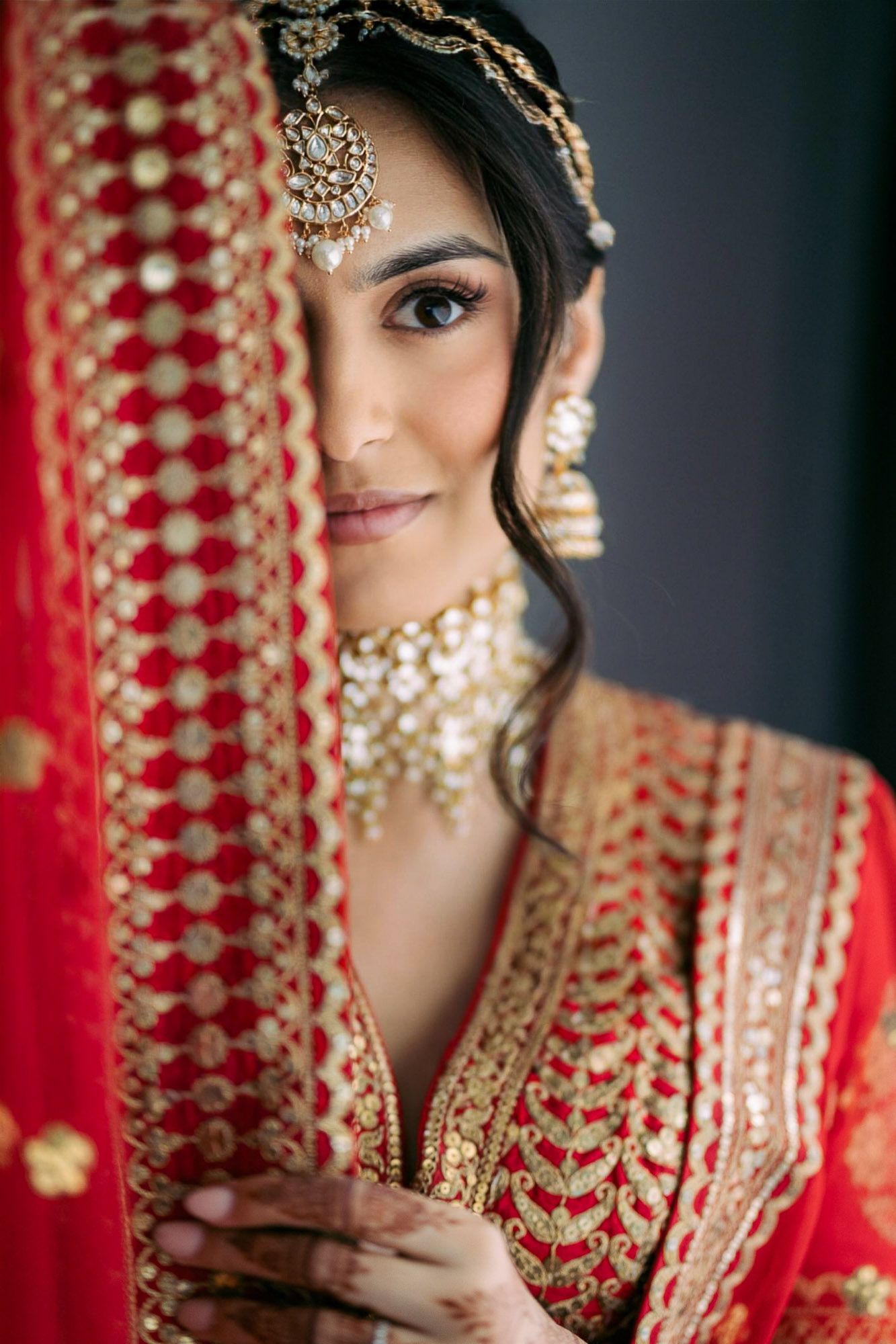 Close-up portrait of a bride wearing traditional Indian bridal jewelry and red attire with ornate gold details.