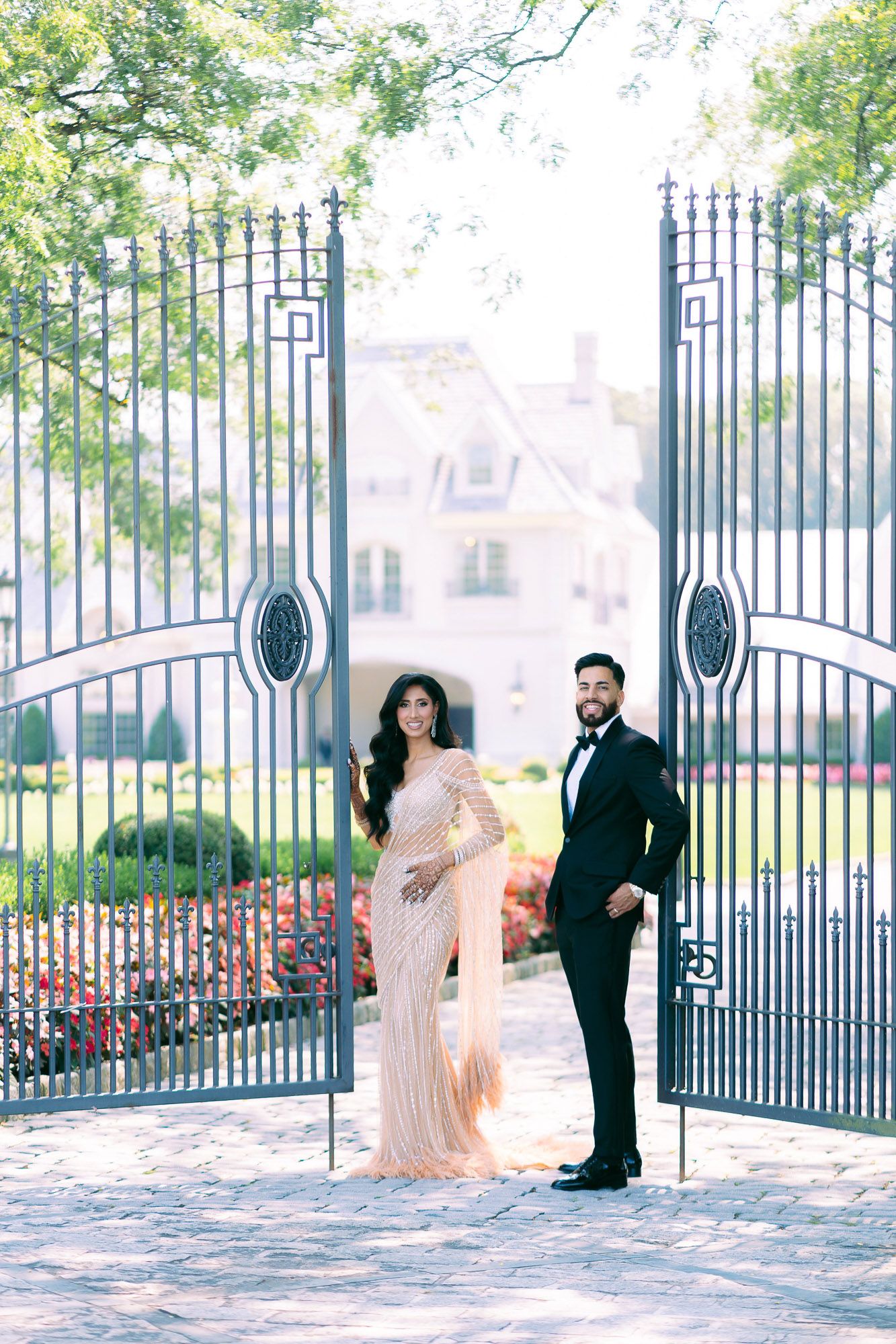 A couple in formal attire poses romantically by an ornate iron gate in a garden setting with soft lighting.