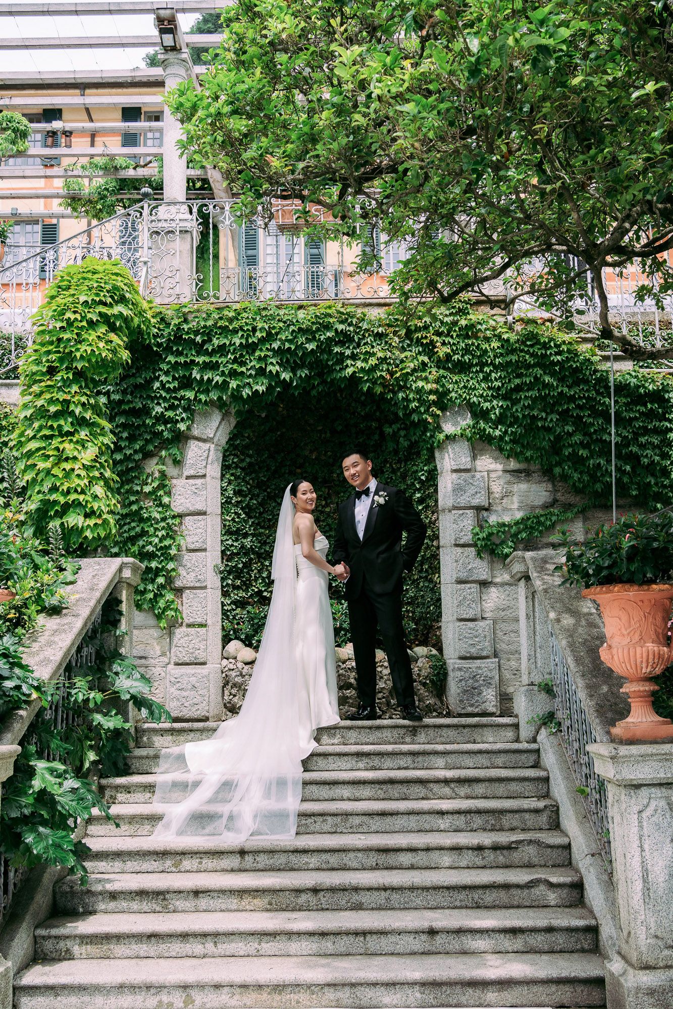 A couple in wedding attire stands on elegant stone steps surrounded by lush greenery and architectural details.