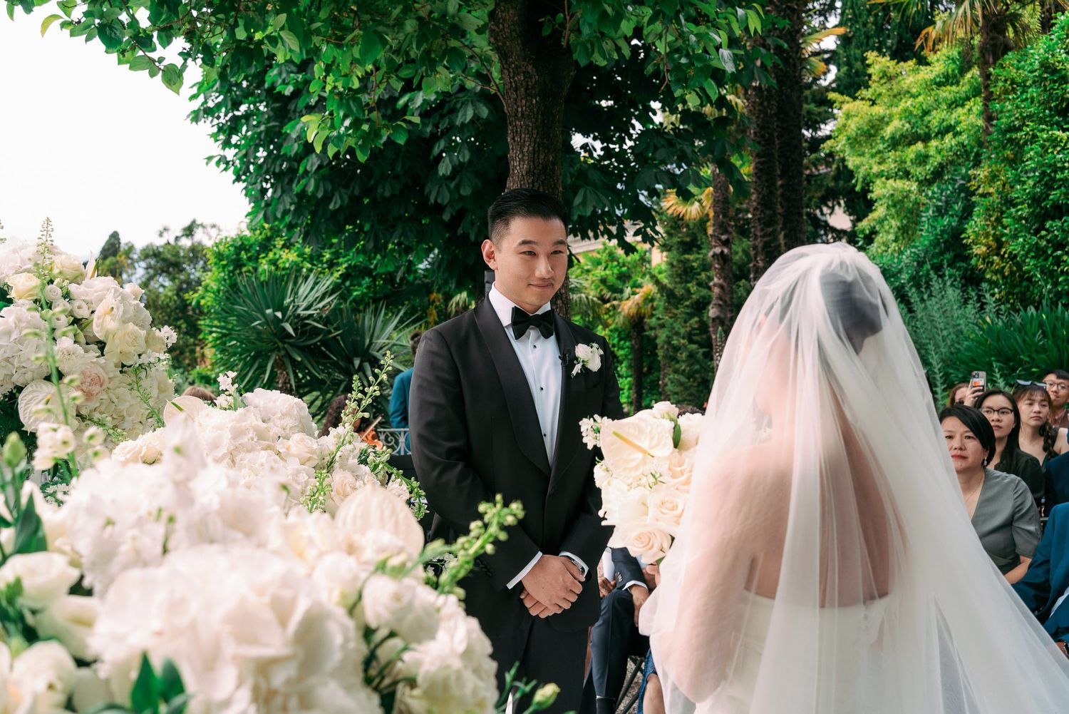 Wedding party walks through a beautiful garden pathway lined with blooming flowers and ornate stone balustrades.
