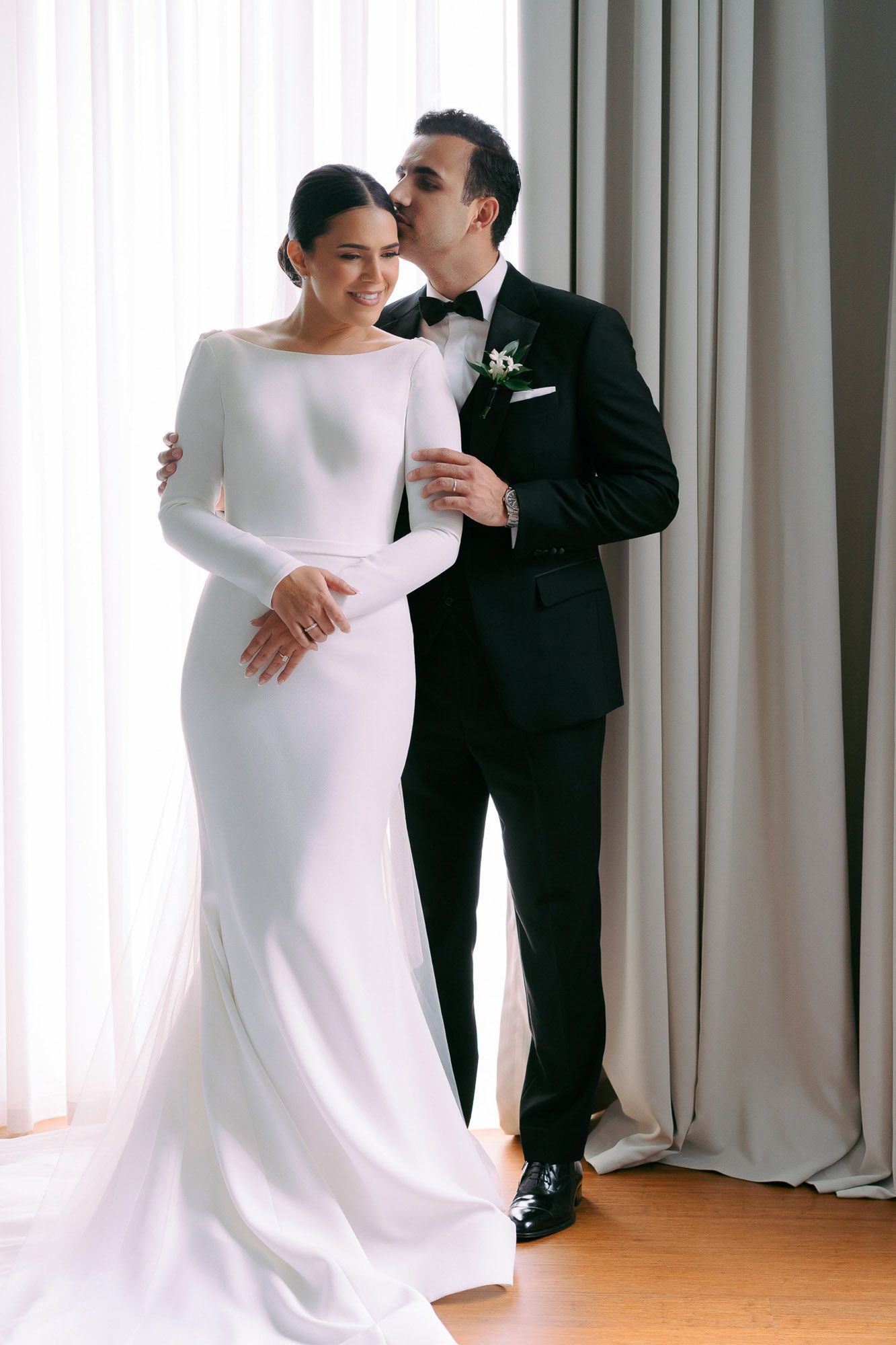 Elegant couple in wedding attire share an intimate moment by white curtains in a modern indoor setting.