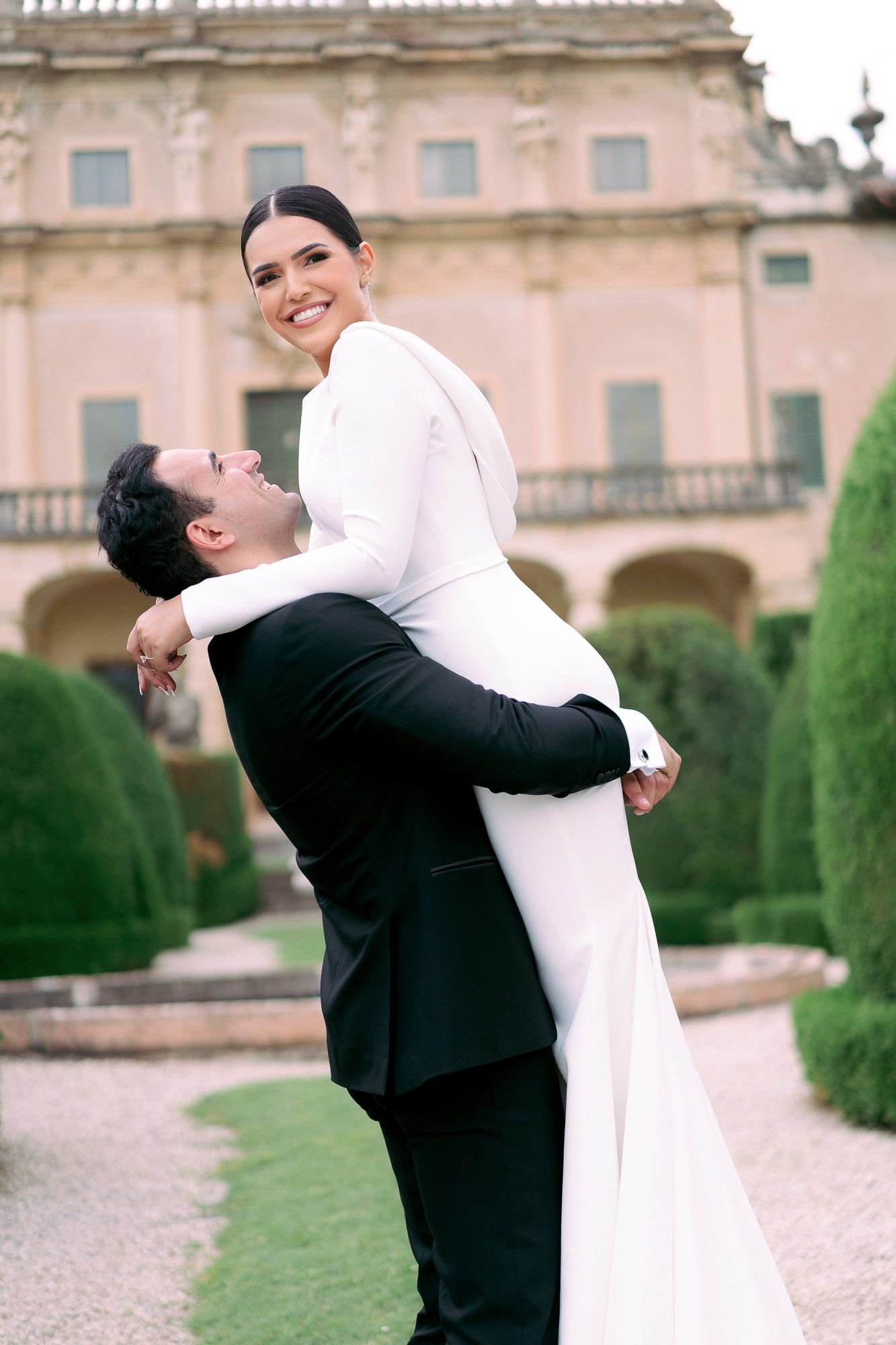 A couple in wedding attire embrace joyfully in front of a grand pink palace with manicured gardens.