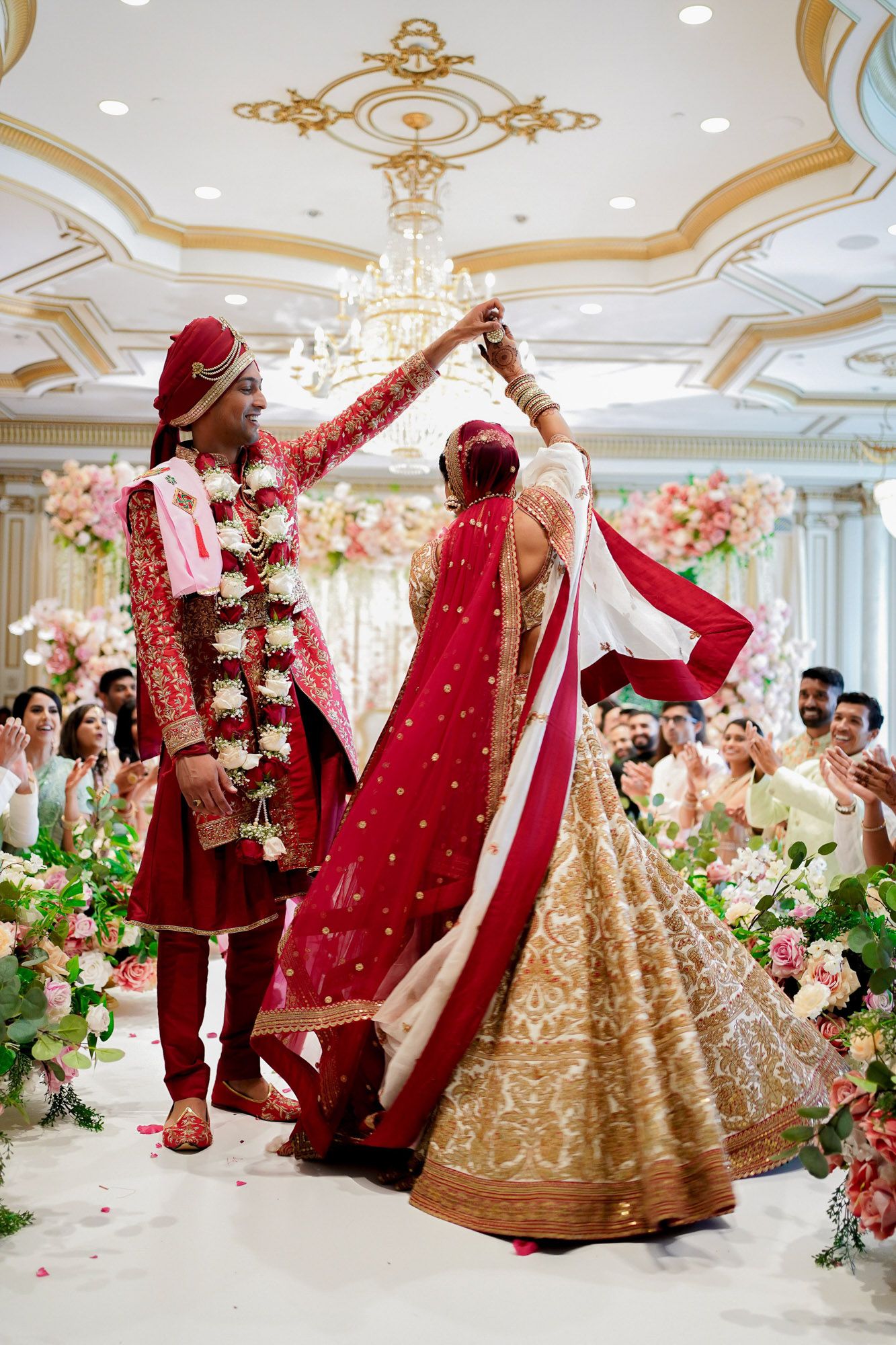 Traditional Indian wedding ceremony with couple in red and gold attire dancing under ornate chandelier at luxury venue.