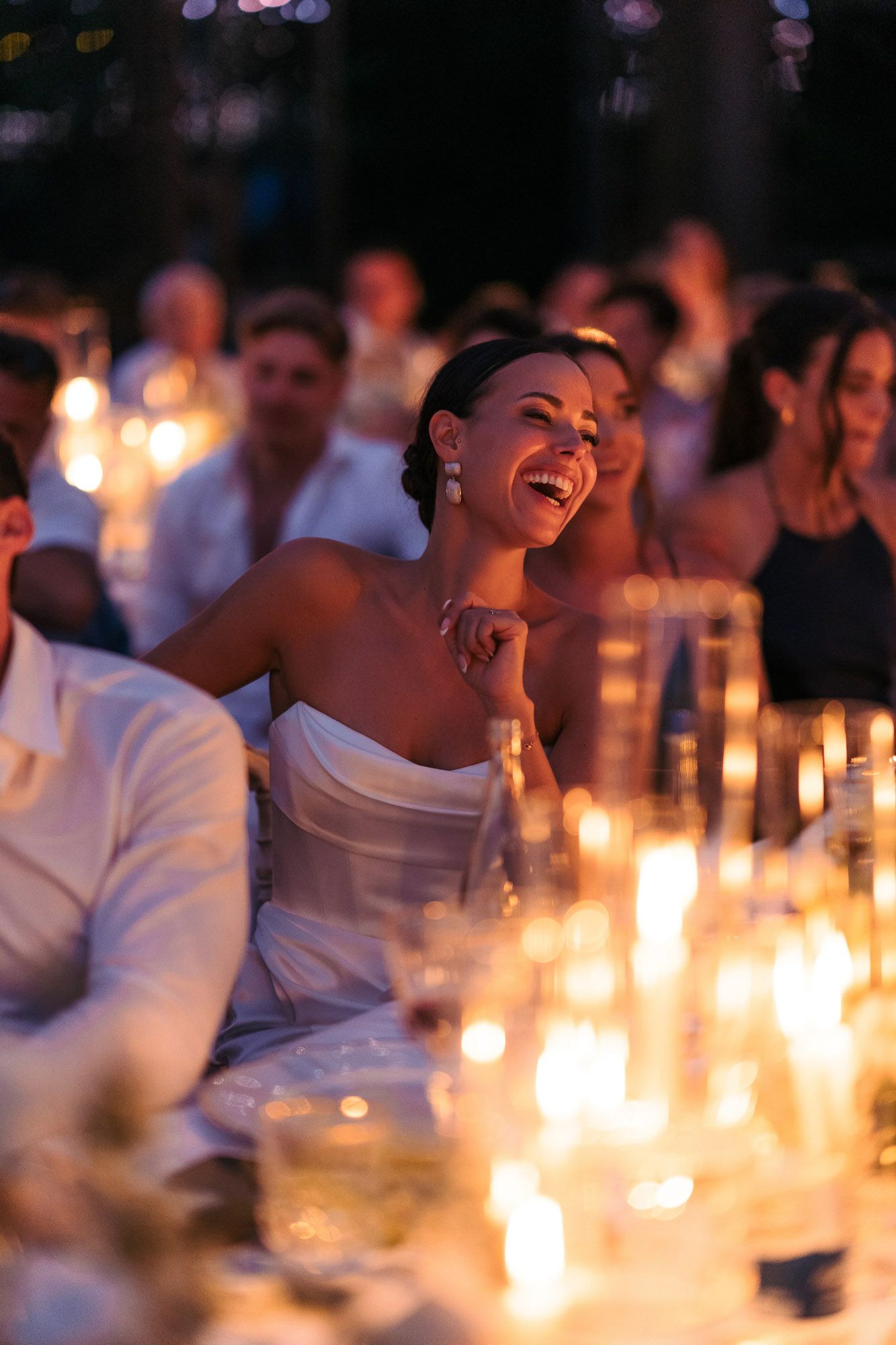 Wedding guests laugh joyfully at a candlelit reception dinner table creating a warm and festive atmosphere.