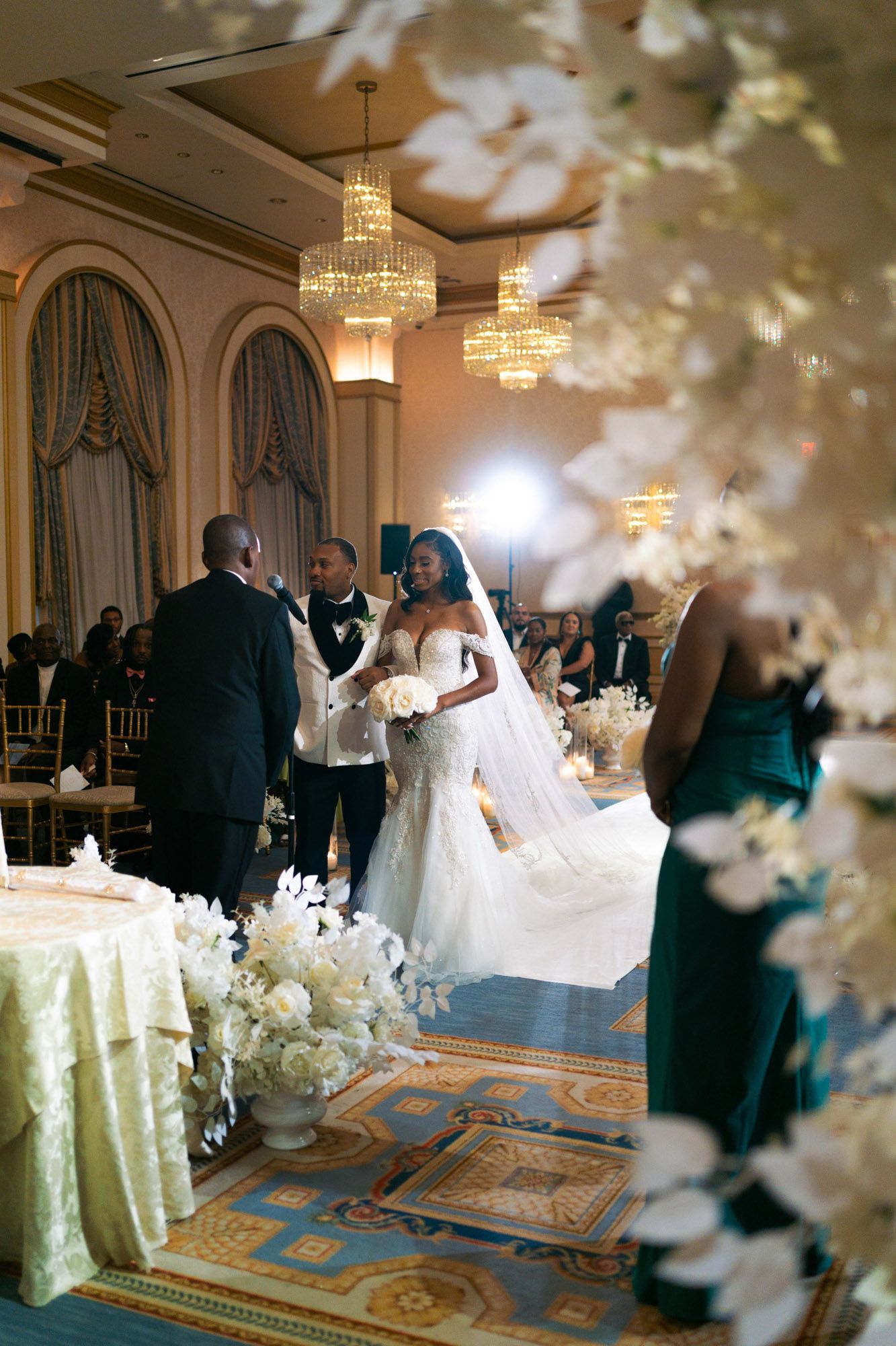 Wedding reception guests dance in an elegant ballroom decorated with crystal chandeliers and white floral arrangements.