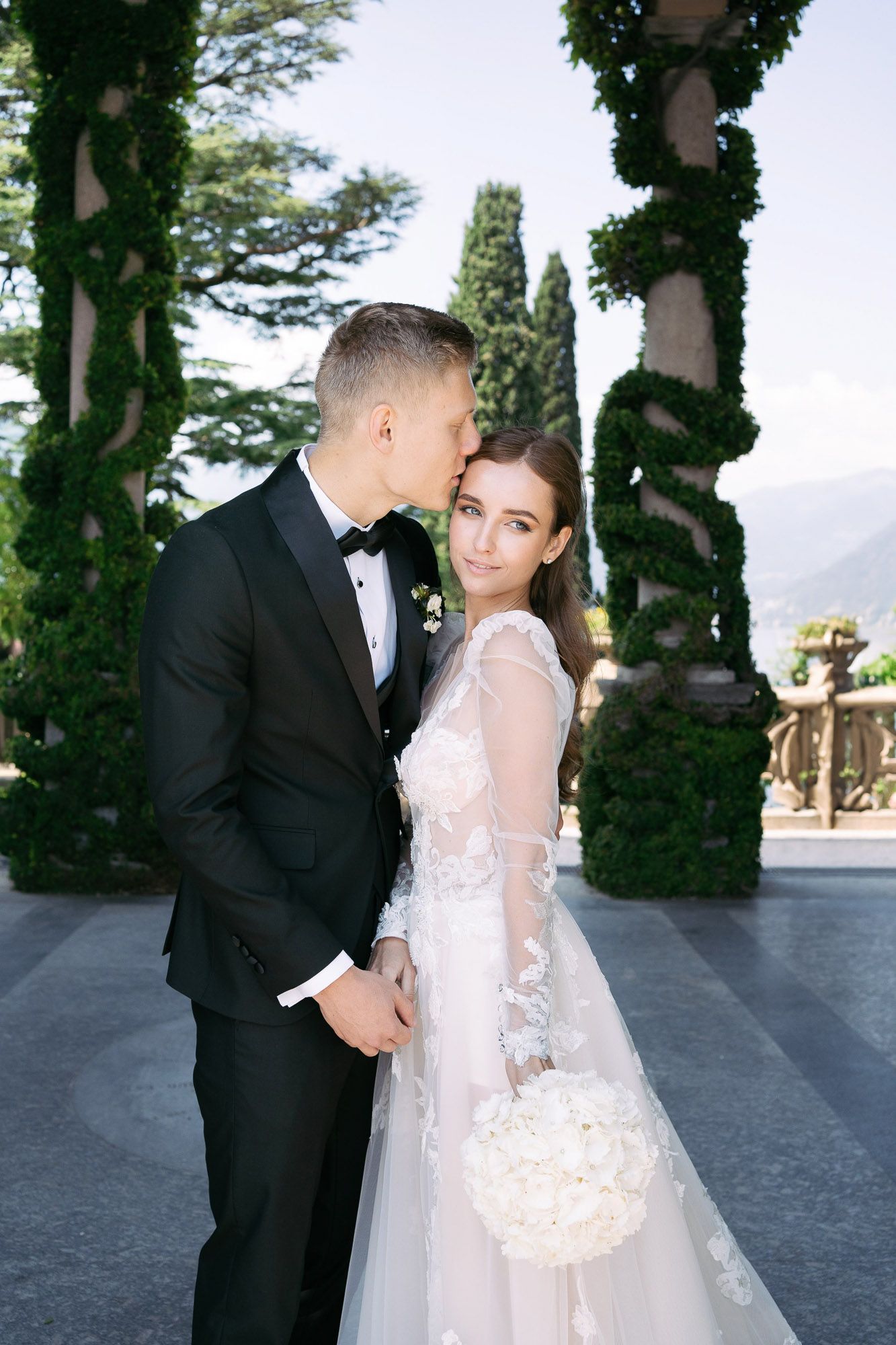 A couple in wedding attire share an intimate moment under ivy-covered columns on a terrace overlooking a scenic view.