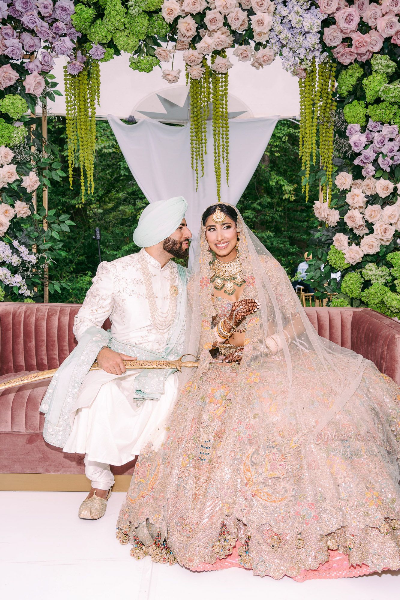 A romantic wedding scene decorated with pink flowers and hanging greenery features a couple in traditional attire.