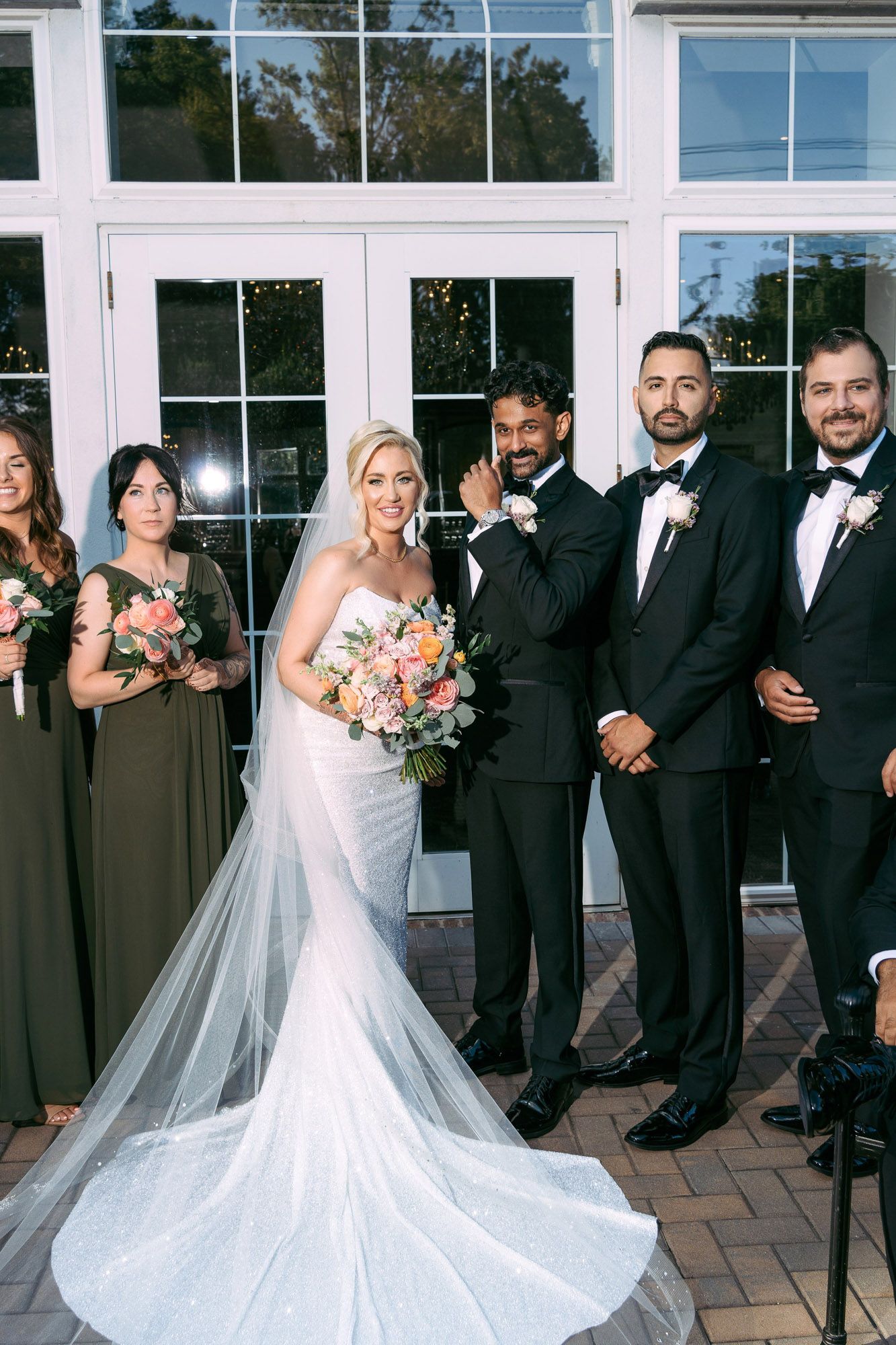 Wedding party in formal attire poses outdoors in front of glass windows with bride in flowing white gown.