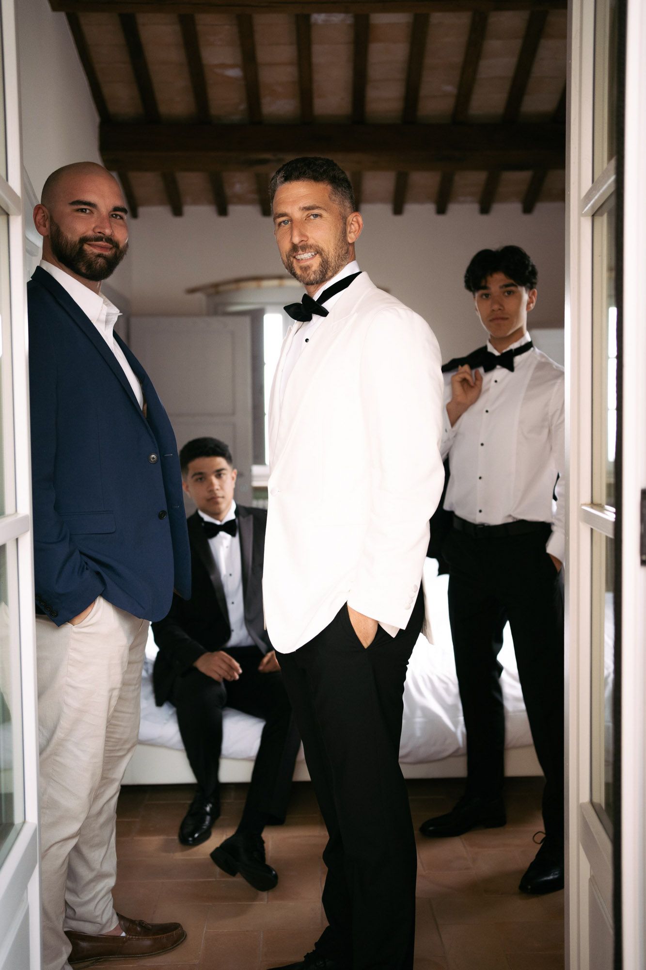 A group of formally dressed people in suits and tuxedos gather in a bright indoor hallway space.