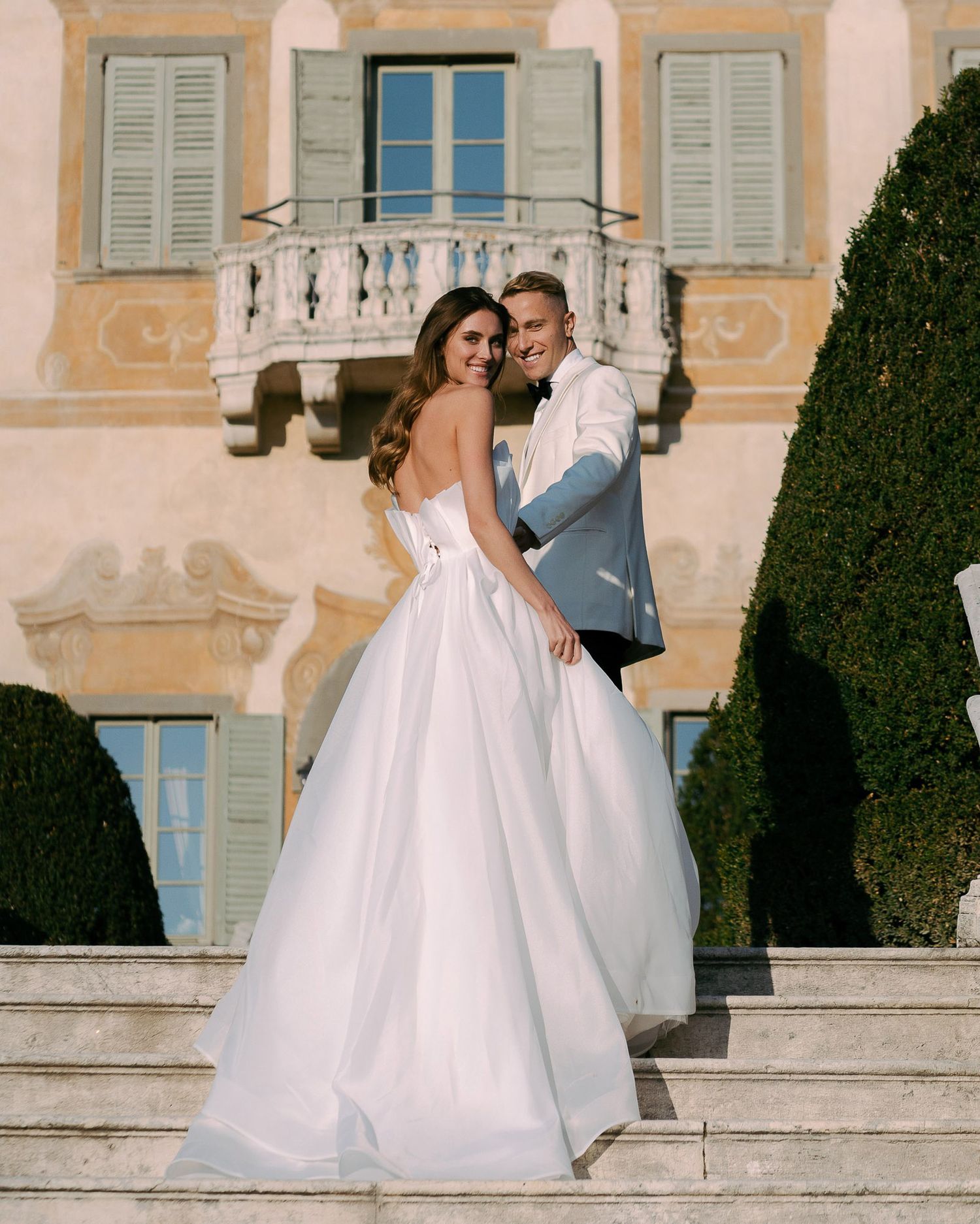 A couple in wedding attire pose on elegant stone steps outside a Mediterranean-style villa with blue shutters.
