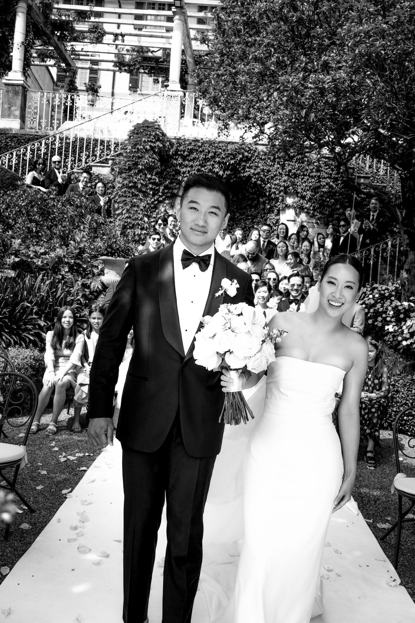 A happy couple poses together on their wedding day in formal attire with a crowd of guests visible in the background.